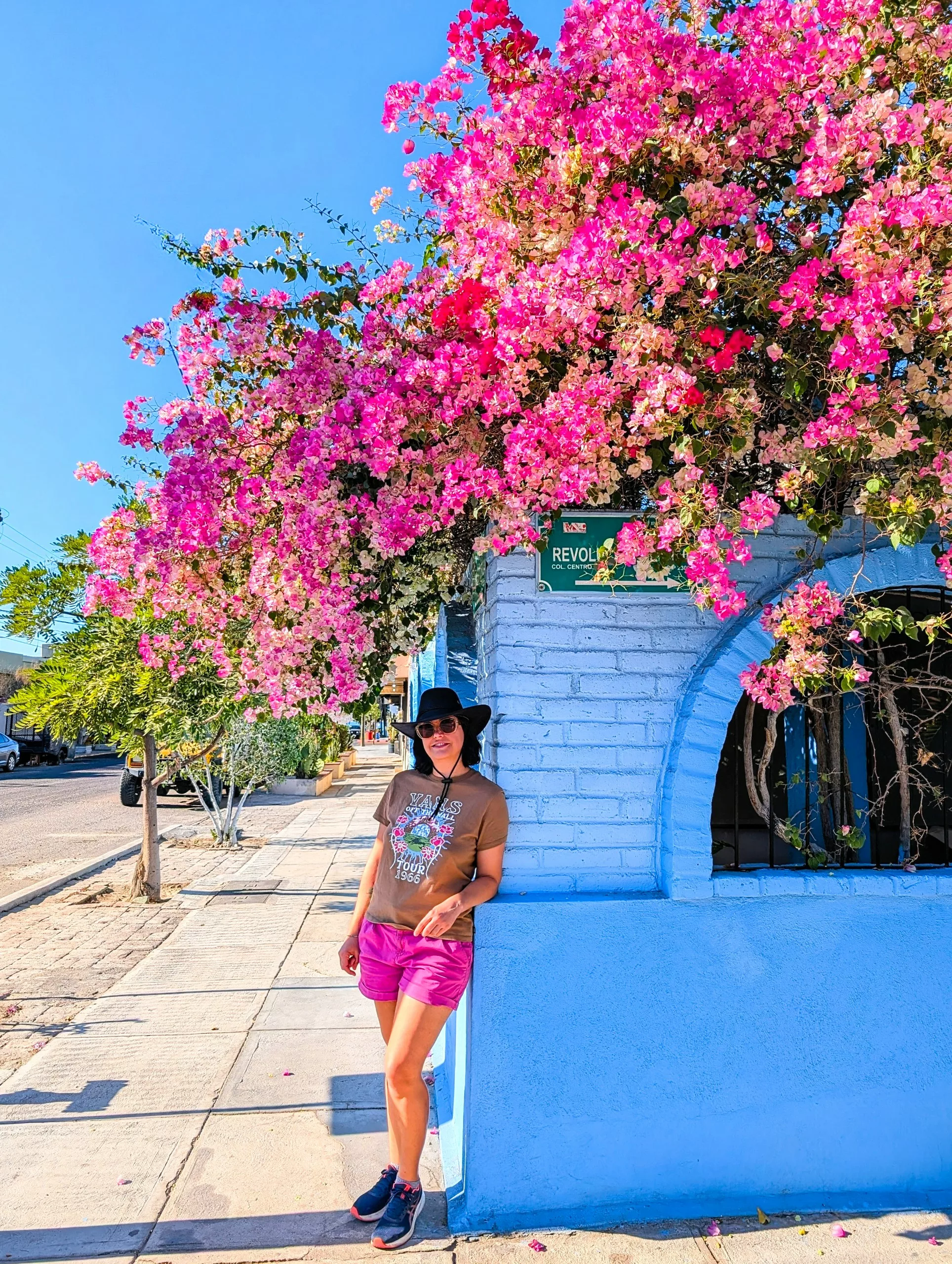 Une femme portant un t-shirt marron, un short rose, des lunettes de soleil et un chapeau noir se tient près d'un mur bleu clair, sous des fleurs roses, sur un trottoir de La Paz, au Mexique.