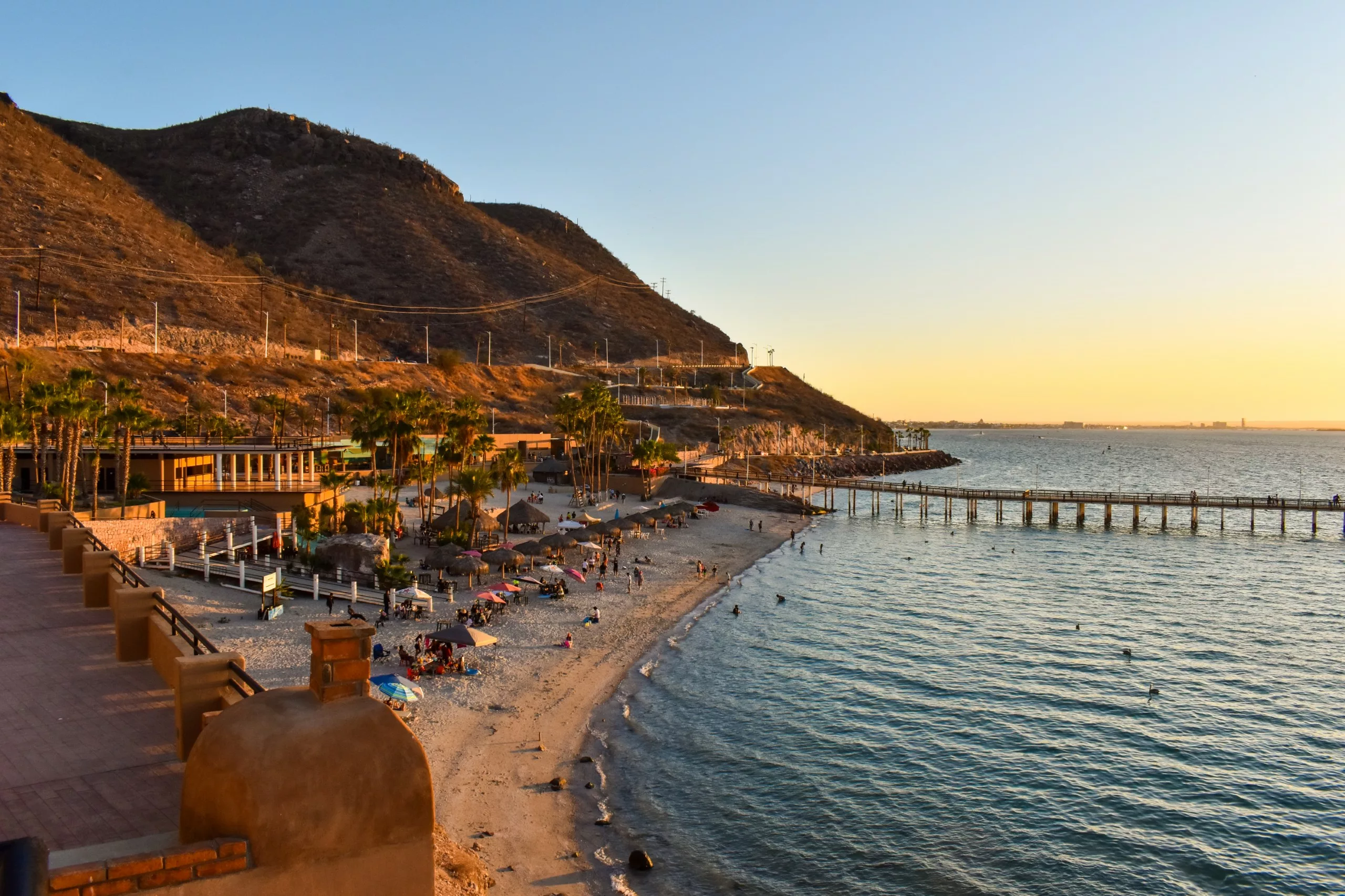 Une plage de sable à La Paz, au Mexique, avec des gens, des parasols et des palmiers à côté d'une eau bleue calme, d'une jetée en bois et de collines au coucher du soleil. Des bâtiments et une route bordent le rivage pittoresque.