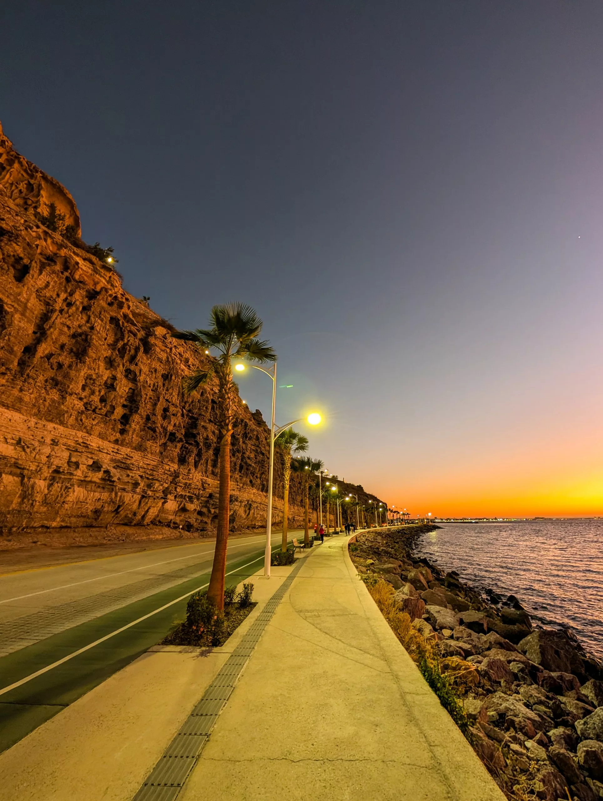 Un sentier pavé et éclairé par des lampadaires longe la côte et une falaise abrupte au coucher du soleil. Le ciel passe du bleu profond à l'orange, la mer est calme sur la droite et des palmiers bordent la promenade.