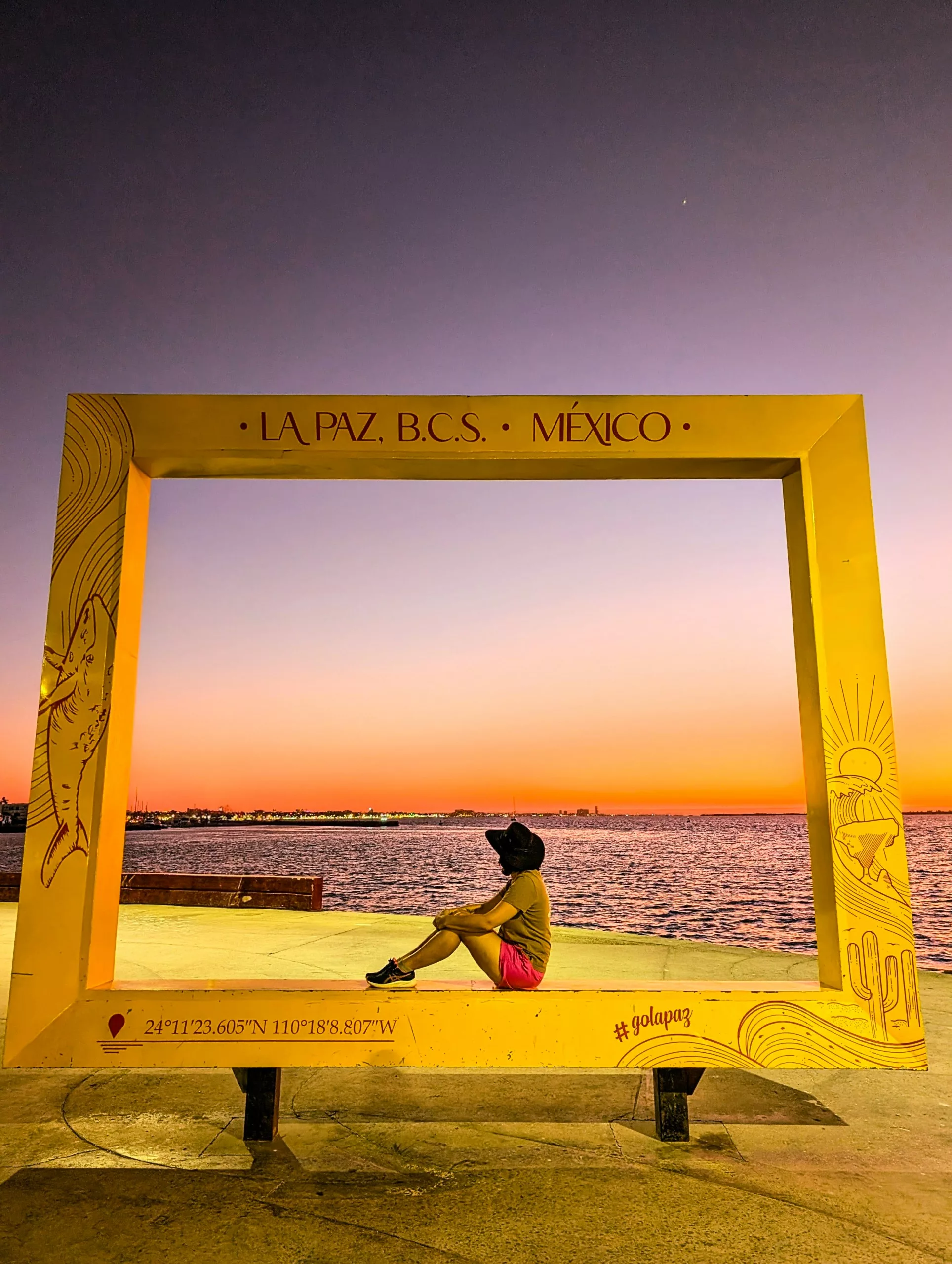 Une personne est assise à l'intérieur d'un grand cadre jaune marqué "La Paz, B.C.S., México", face à l'océan au coucher du soleil. Le ciel s'illumine d'orange et de violet ; les coordonnées GPS sont imprimées sur le cadre.