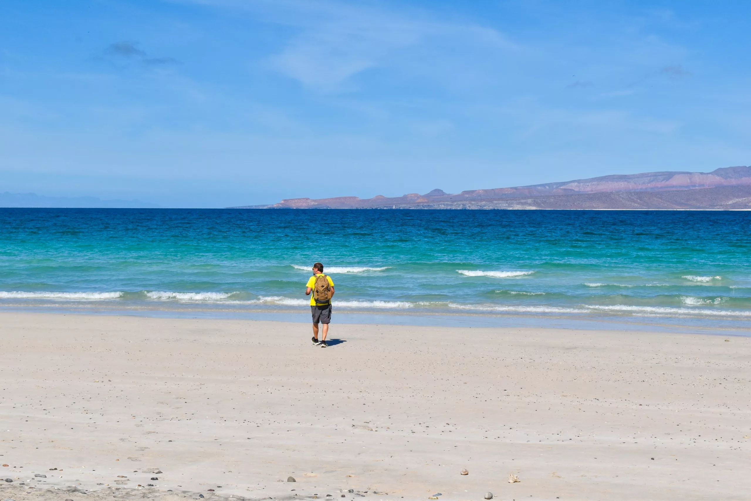 Un homme portant un-shirt jaune et un sac à dos se tient sur une plage de sable face à l'océan, avec de douces vagues et des montagnes lointaines sous un ciel bleu.