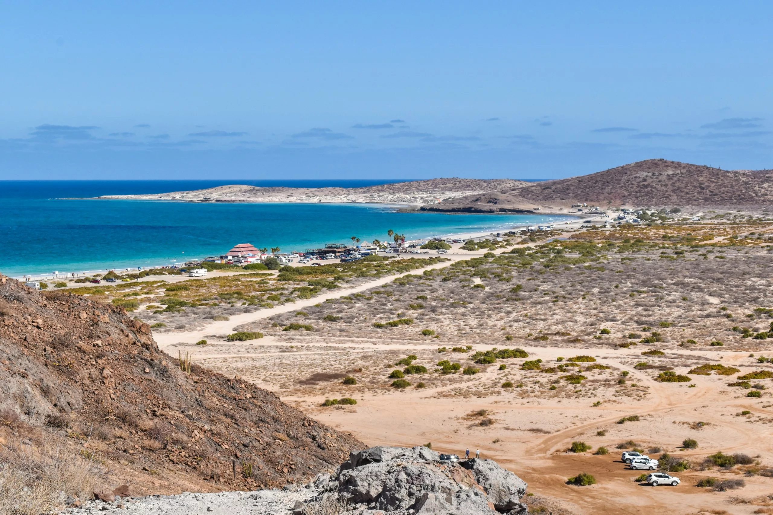 Paysage côtier avec un terrain sablonneux, une végétation basse et quelques véhicules éparpillés. Le littoral s'incurve le long de l'eau bleue de l'océan, avec de petites collines au loin, sous un ciel clair.