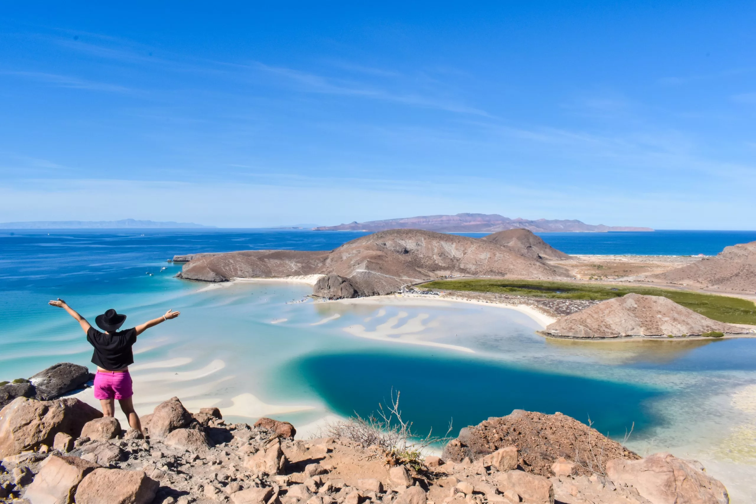 Une personne se tient debout sur un terrain rocheux, les bras levés, surplombant une eau bleue, plages de sable et îles lointaines sous un ciel bleu limpide.