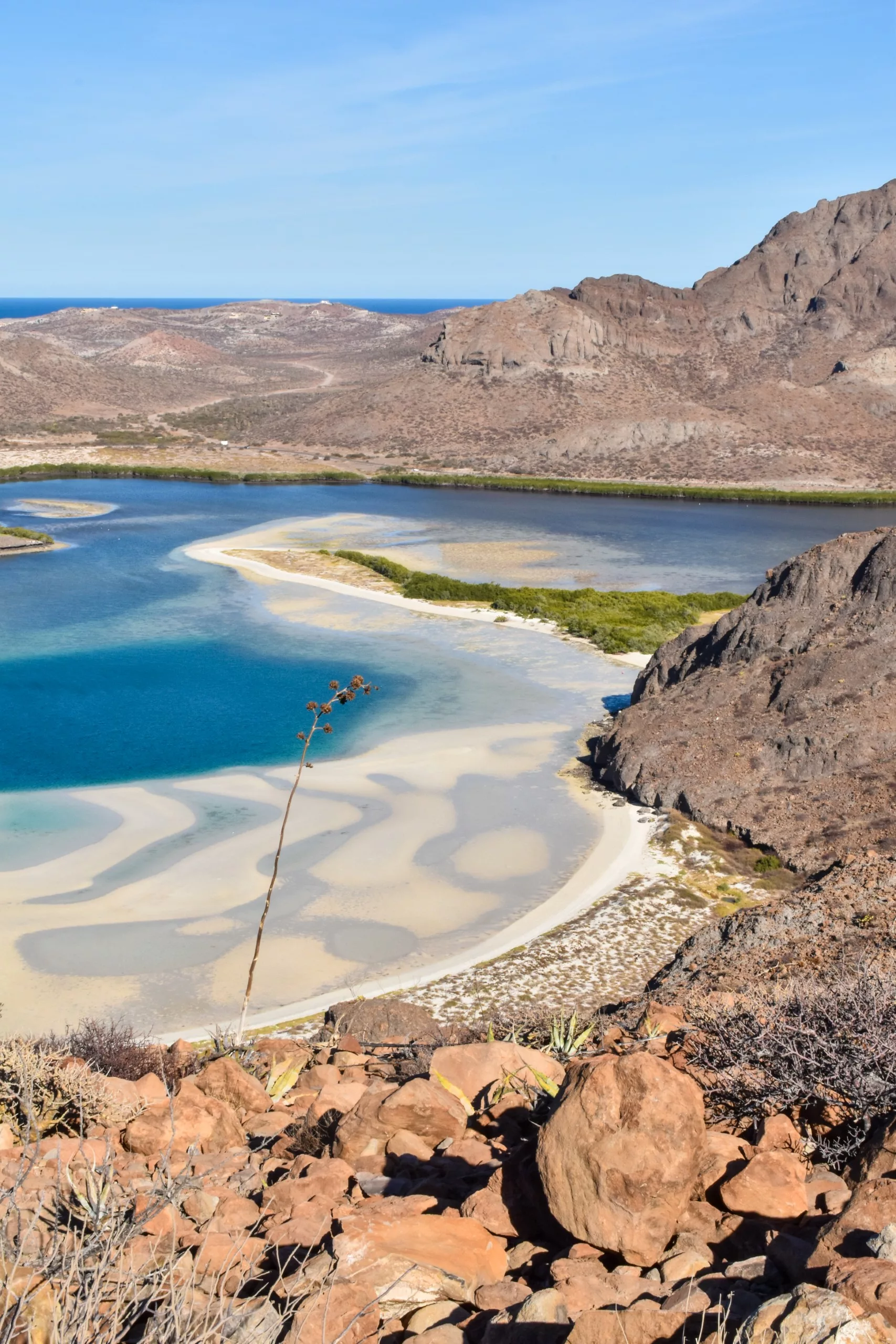 Une lagune bleue avec des bancs de sable et des arbustes verts serpente à travers un paysage rocheux et aride. Des montagnes brunes entourent l'eau et des plantes sèches sont visibles au premier plan.