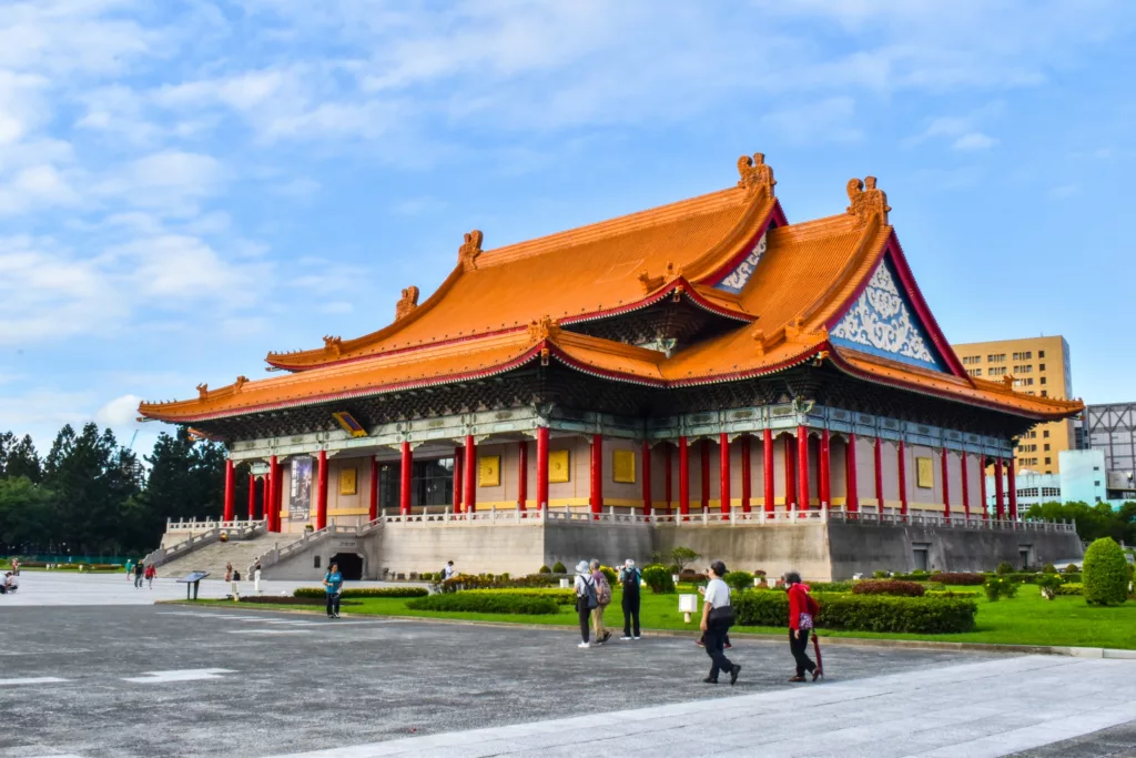 Un grand bâtiment chinois traditionnel avec un toit rouge et orange, entouré de gens qui marchent sur une place à Taipei