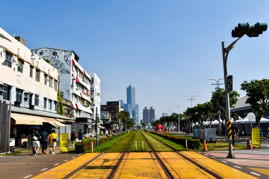 Une rue de la ville avec des voies de tramway est bordée de bâtiments et d'arbres Les gens se promènent sur les trottoirs tandis qu'un gratte-ciel s'élève à l'arrière-plan sous un ciel bleu limpide.