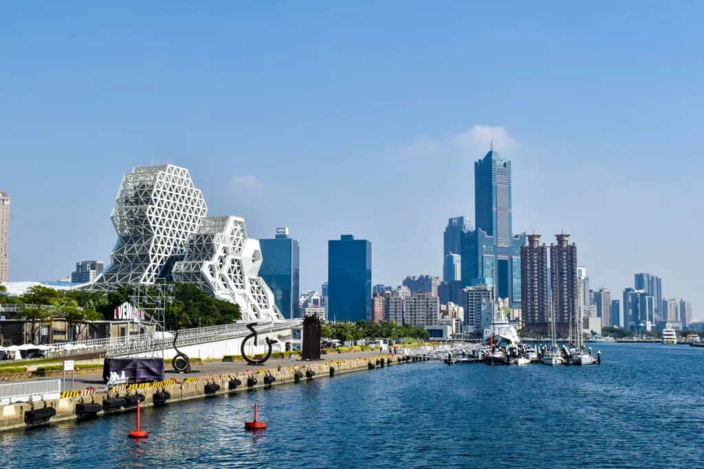 Vue d'un paysage urbain au bord de l'eau à Taïwan, avec des bâtiments modernes - dont une structure géométrique blanche à gauche - et des tours d'habitation à l'arrière-plan, à côté d'une jetée avec des bateaux amarrés le long de l'eau.
