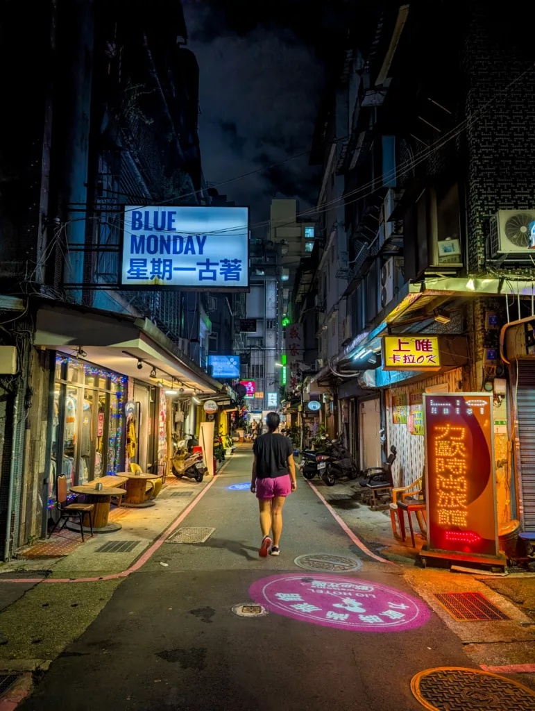 Une personne en short marche dans une ruelle urbaine étroite et très éclairée la nuit. Des enseignes au néon en anglais et en chinois illuminent la rue.