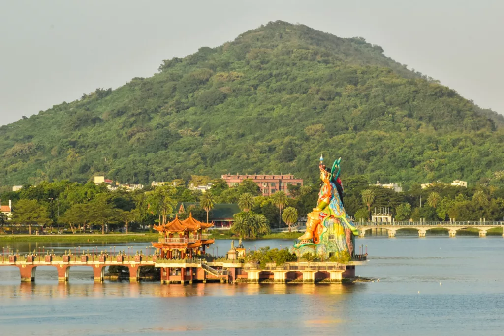 Une scène au bord du lac avec un pavillon chinois traditionnel sur une jetée, une grande statue colorée et des collines verdoyantes.