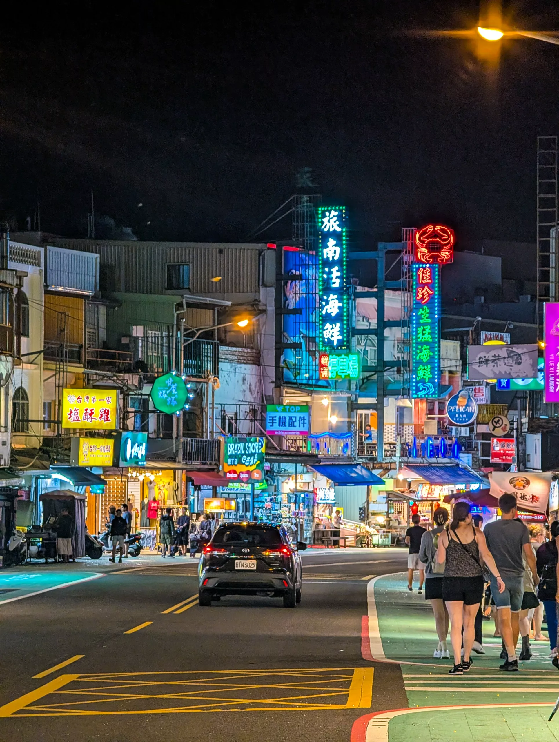 Une rue animée la nuit, avec des néons en chinois, des lumières vives, des scooters garés et des gens qui marchent sur le trottoir.