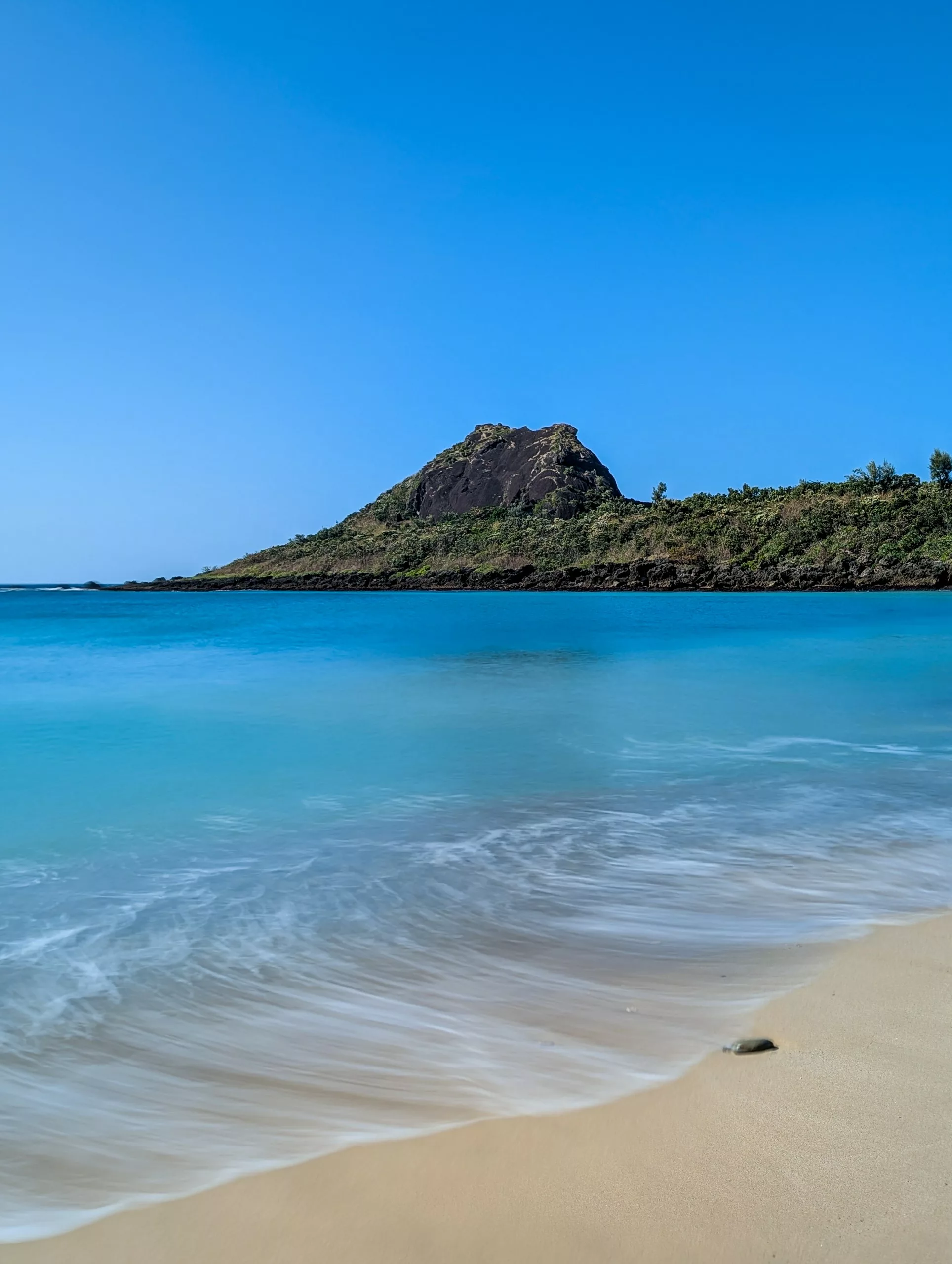 Une plage de sable avec de douces vagues au premier plan, une eau bleue claire et une colline verte avec un pic rocheux.