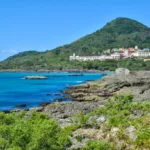 Une côte rocheuse avec une eau turquoise, de la verdure au premier plan et un groupe de bâtiments au pied d'une colline couverte d'arbres sous un ciel bleu clair