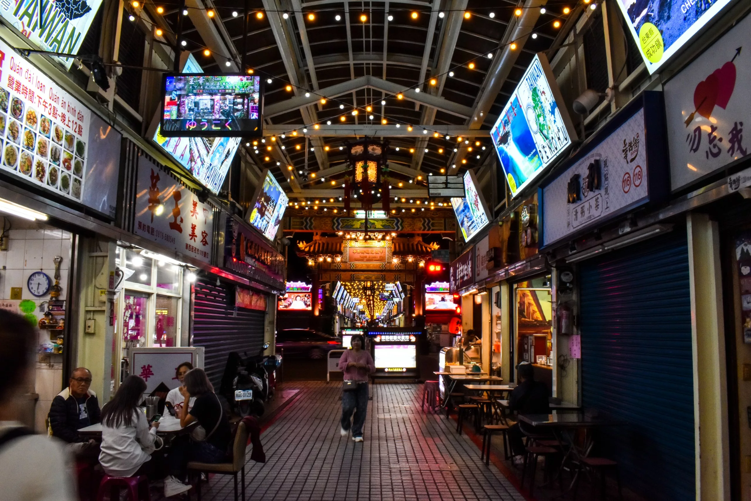 Une allée de marché couverte la nuit, avec des enseignes lumineuses et des guirlandes électriques au-dessus de la tête