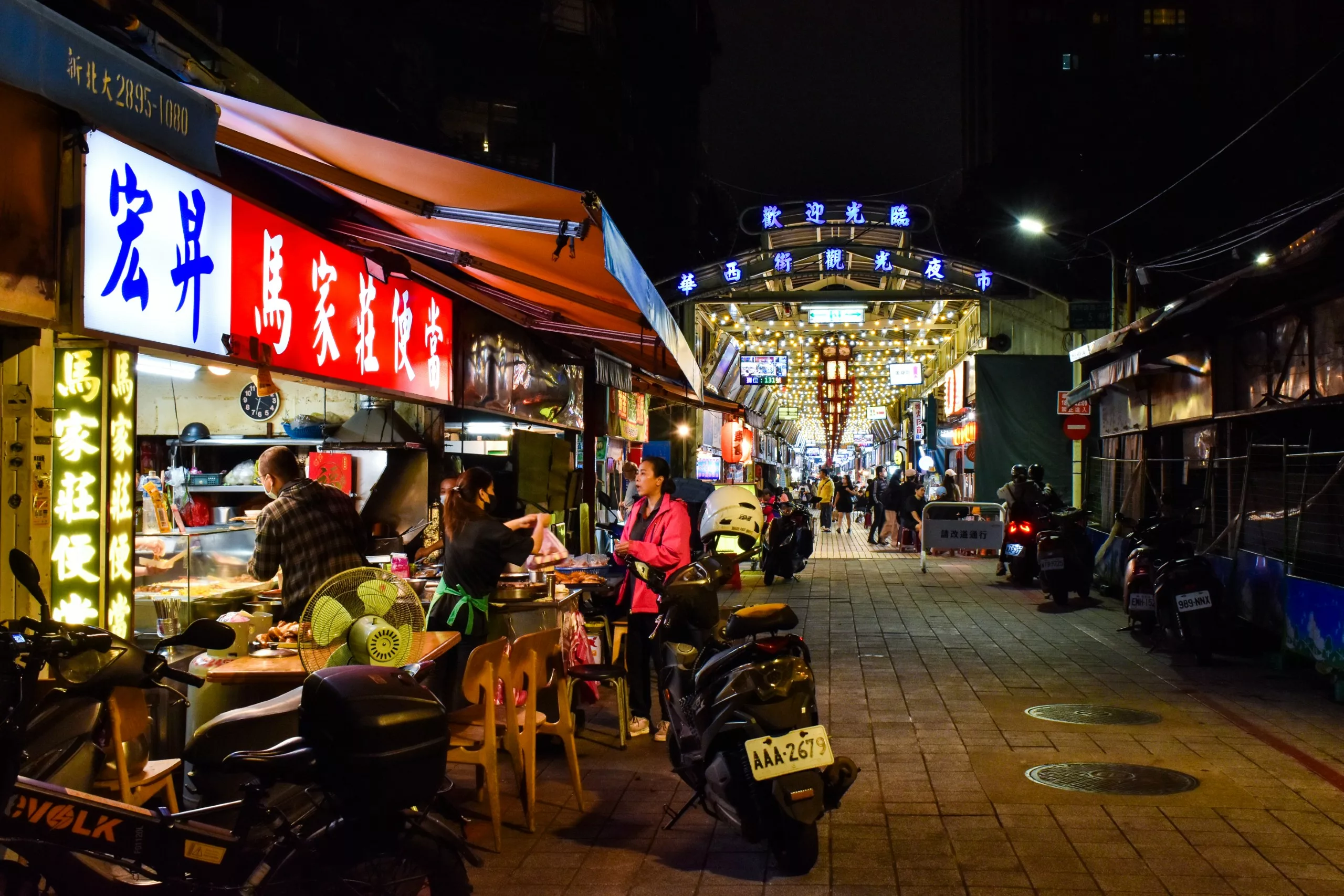 Une scène de marché nocturne avec des gens qui mangent sur des étals de nourriture en plein air, des panneaux lumineux en chinois, des scooters garés et une entrée de marché illuminée à l'arrière-plan.
