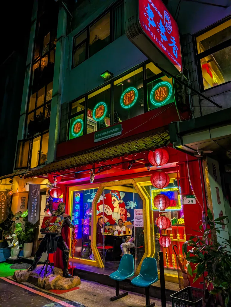 Un restaurant très éclairé la nuit à Taipei, avec des enseignes au néon, des lanternes rouges, une grande vitrine illustrée, une sculpture de Thor et des chaises bleues à l'extérieur 