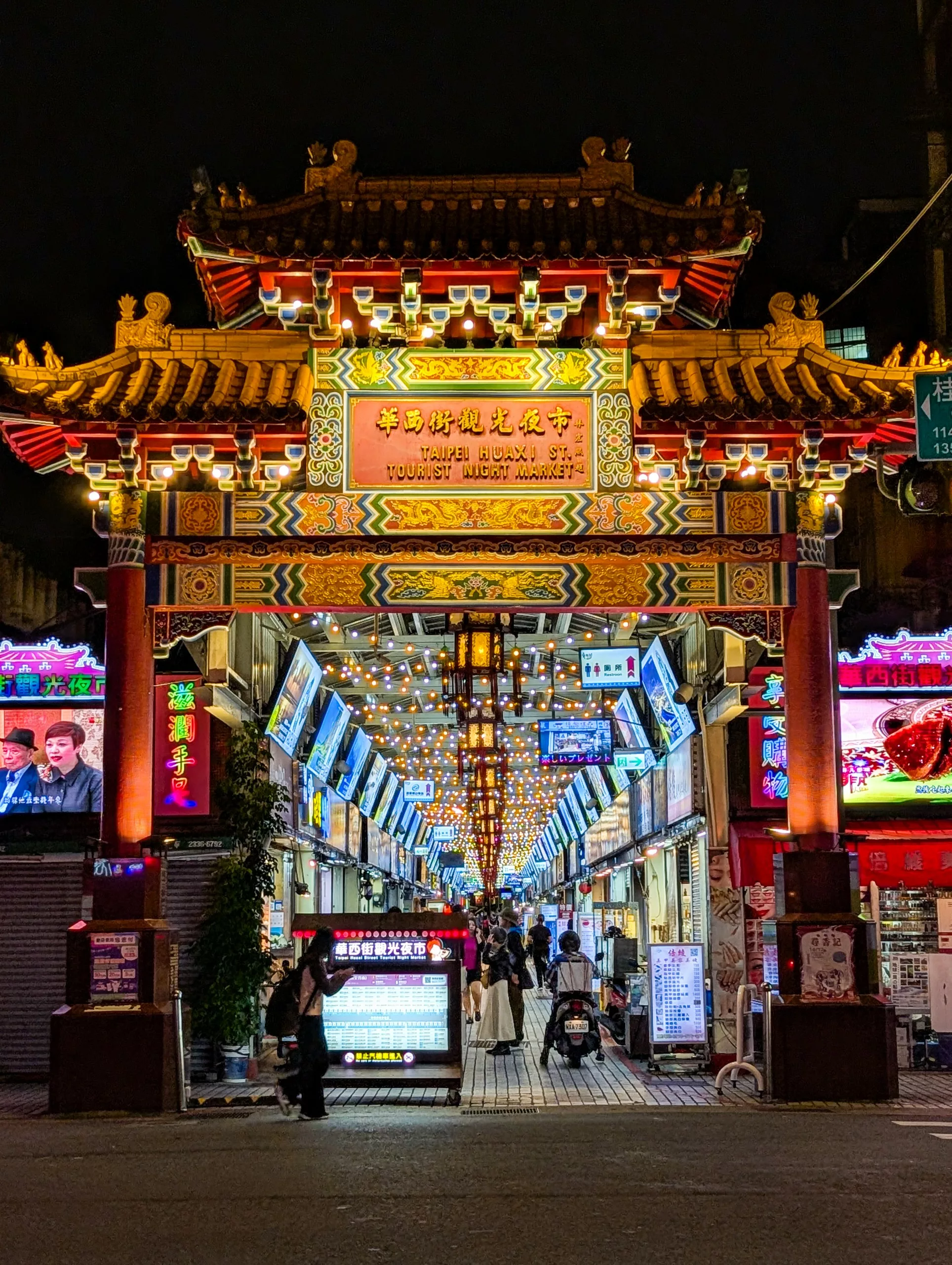 Une porte chinoise traditionnelle illuminée marque l'entrée d'un marché de nuit. Des enseignes au néon et des lanternes éclairent l'allée animée où les gens se promènent la nuit.