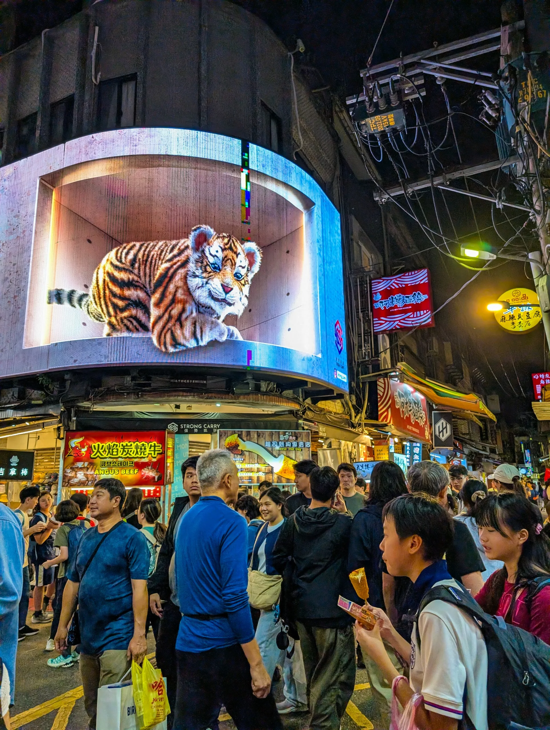 Un marché de nuit bondé à Taipei avec des gens marchant sous un grand panneau d'affichage numérique incurvé affichant un tigre animé en 3D