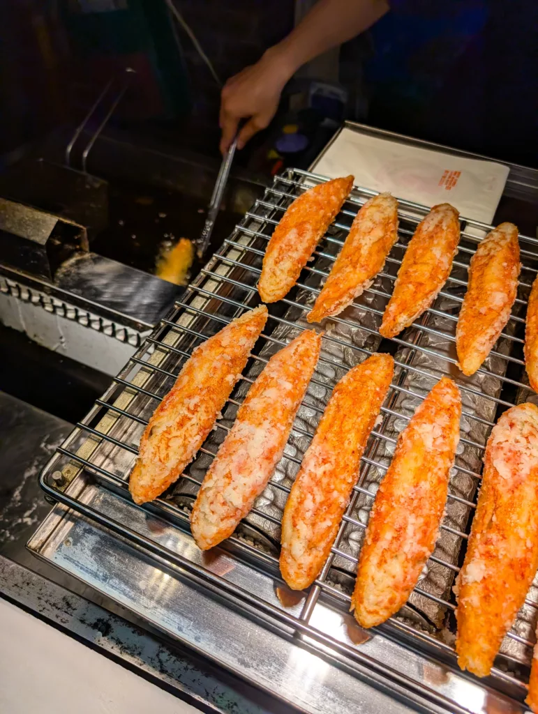 Plusieurs filets de crabes panés sont disposés sur une grille métallique, et une personne à l'arrière-plan fait frire un autre filet dans de l'huile chaude à l'aide de pinces métalliques