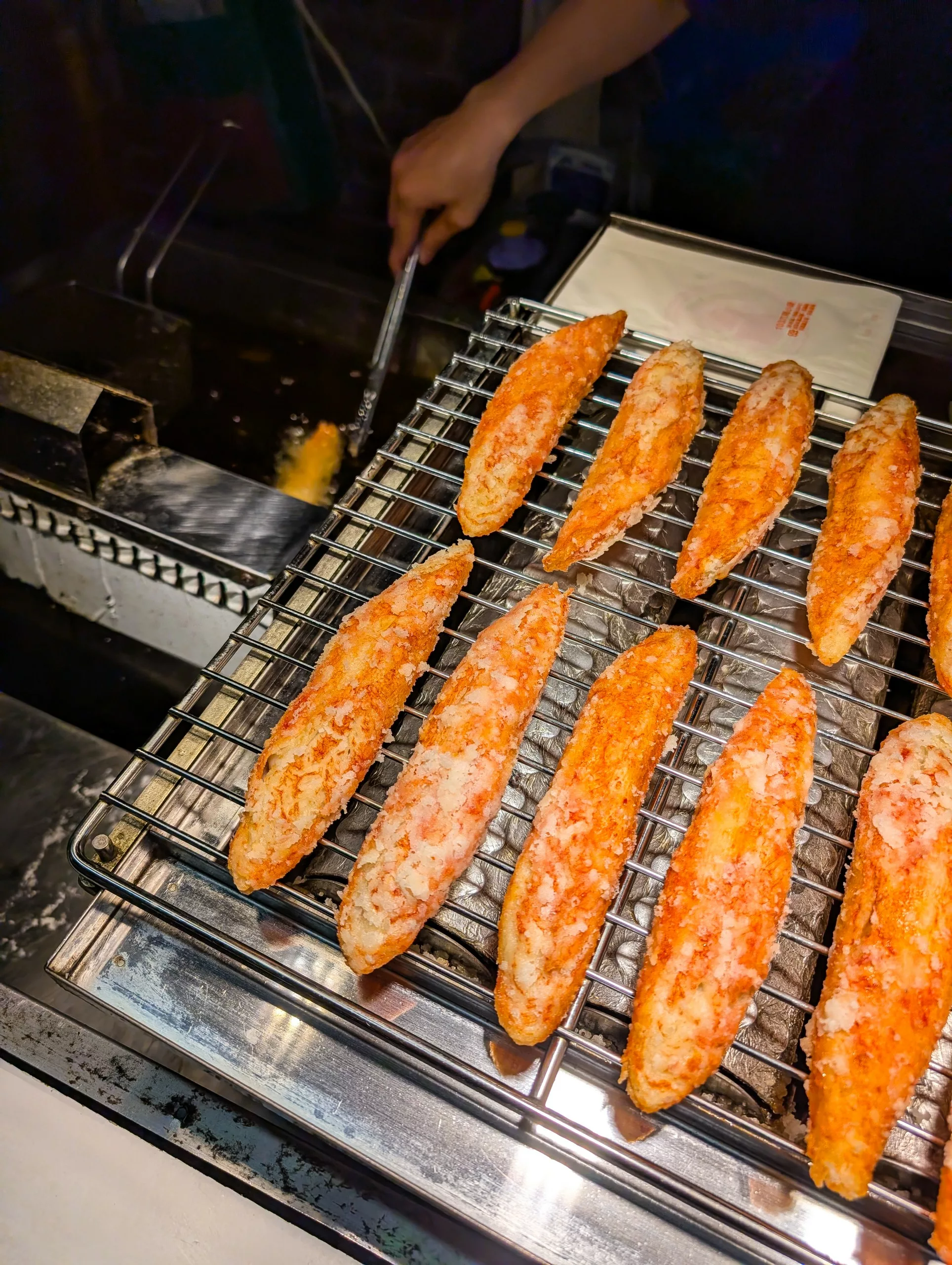 Plusieurs filets de crabes panés sont disposés sur une grille métallique, et une personne à l'arrière-plan fait frire un autre filet dans de l'huile chaude à l'aide de pinces métalliques