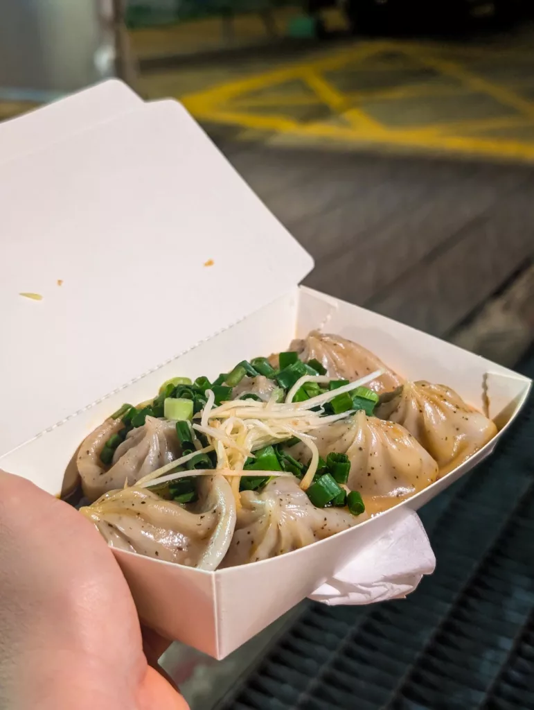 Une main tient une boîte blanche ouverte contenant six ravioles à la vapeur garnies d'oignons verts et de gingembre. 