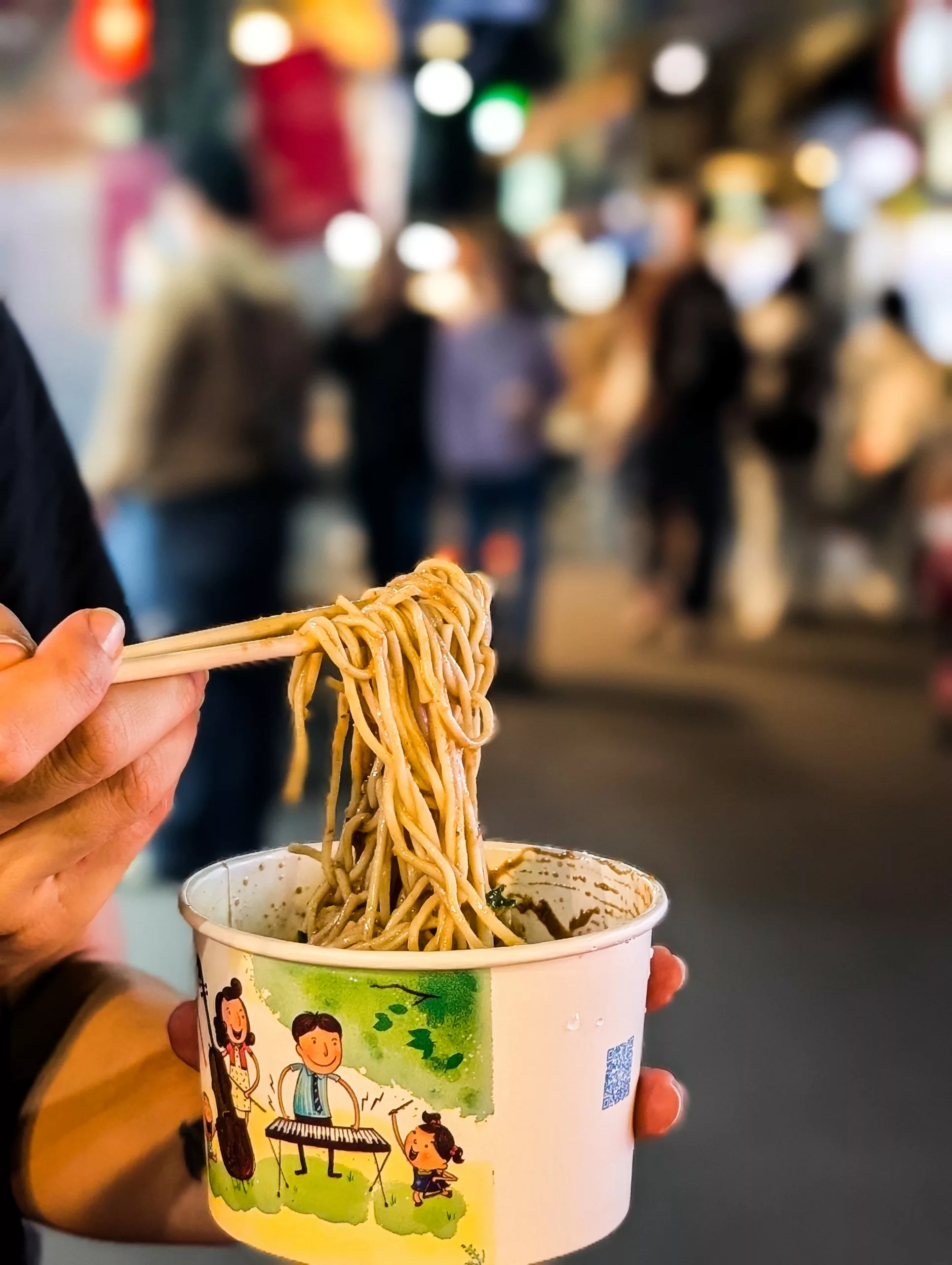 Une personne tenant une tasse de nouilles avec des baguettes dans une main. Le gobelet est orné d'illustrations de bandes dessinées. L'arrière-plan flou, animé par des personnes et des lumières, laisse entrevoir que faire à Taipei dans un marché de rue animé.
