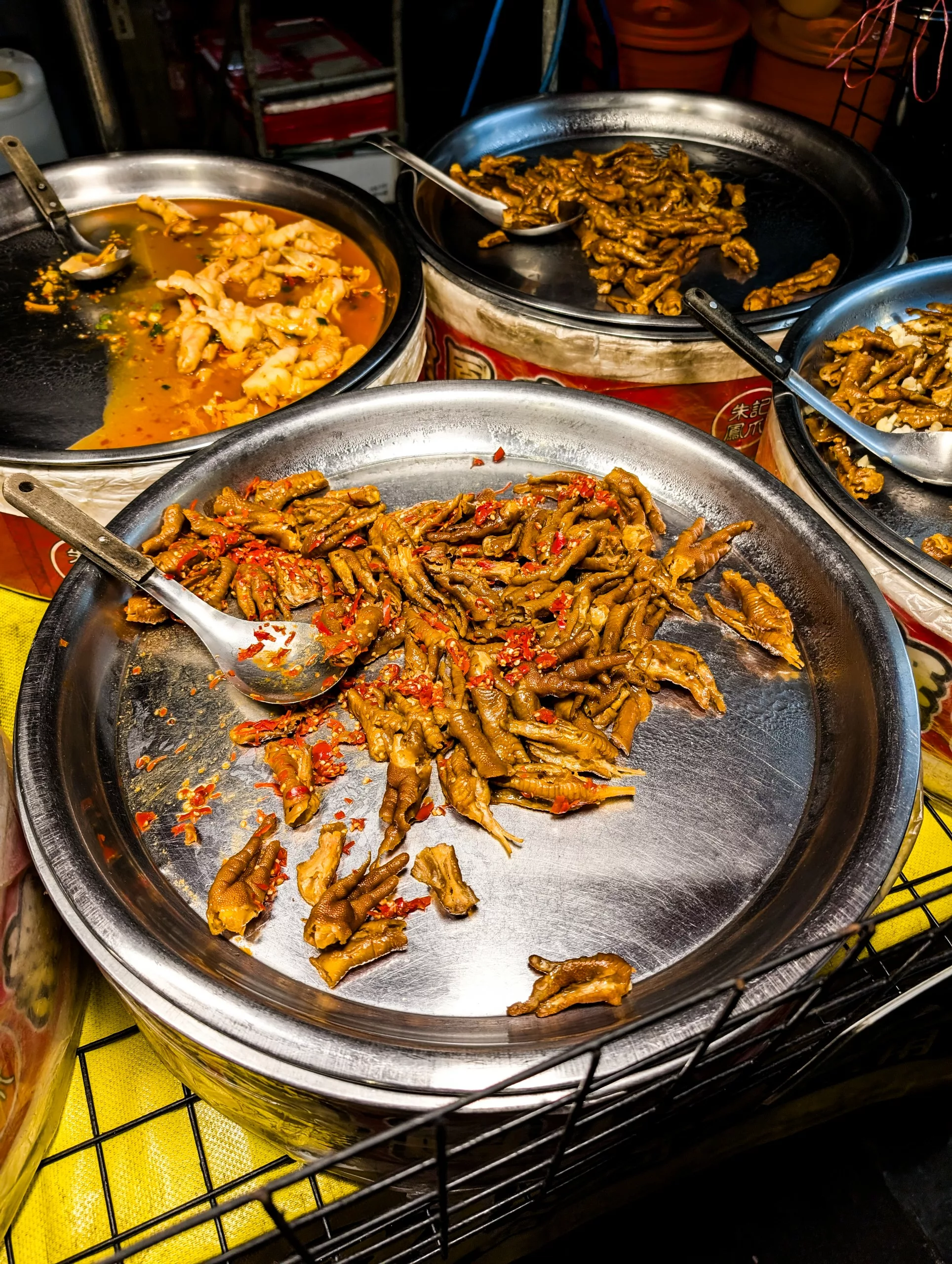 Un plateau métallique rempli de pattes de poulet cuites et assaisonnées de piment rouge, posé sur une surface jaune dans un stand de nourriture. Des cuillères de service sont posées sur chaque plateau.