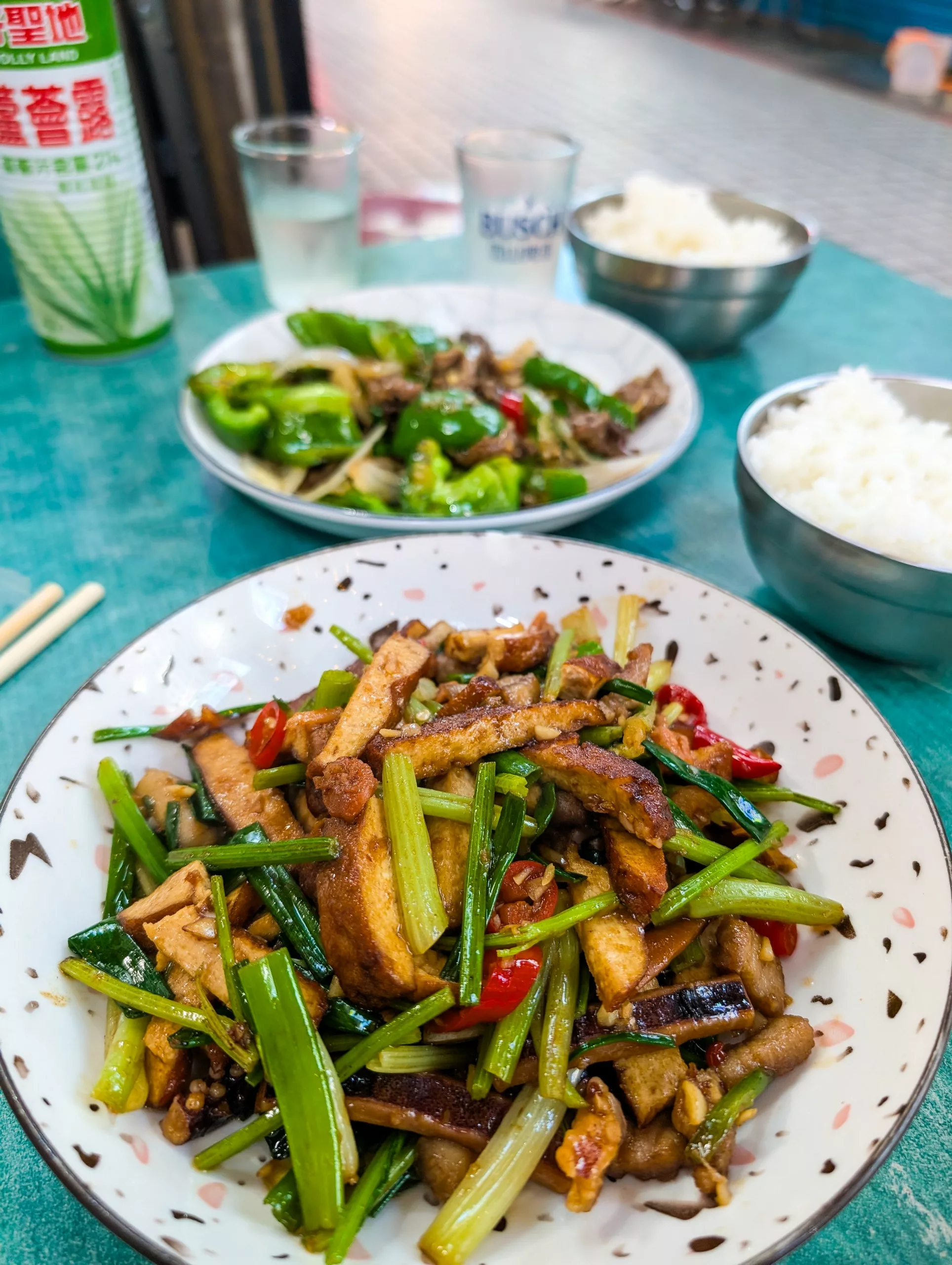 Une table avec deux assiettes de légumes et de tofu sautés, deux bols de riz blanc et des boissons, installée en plein air 