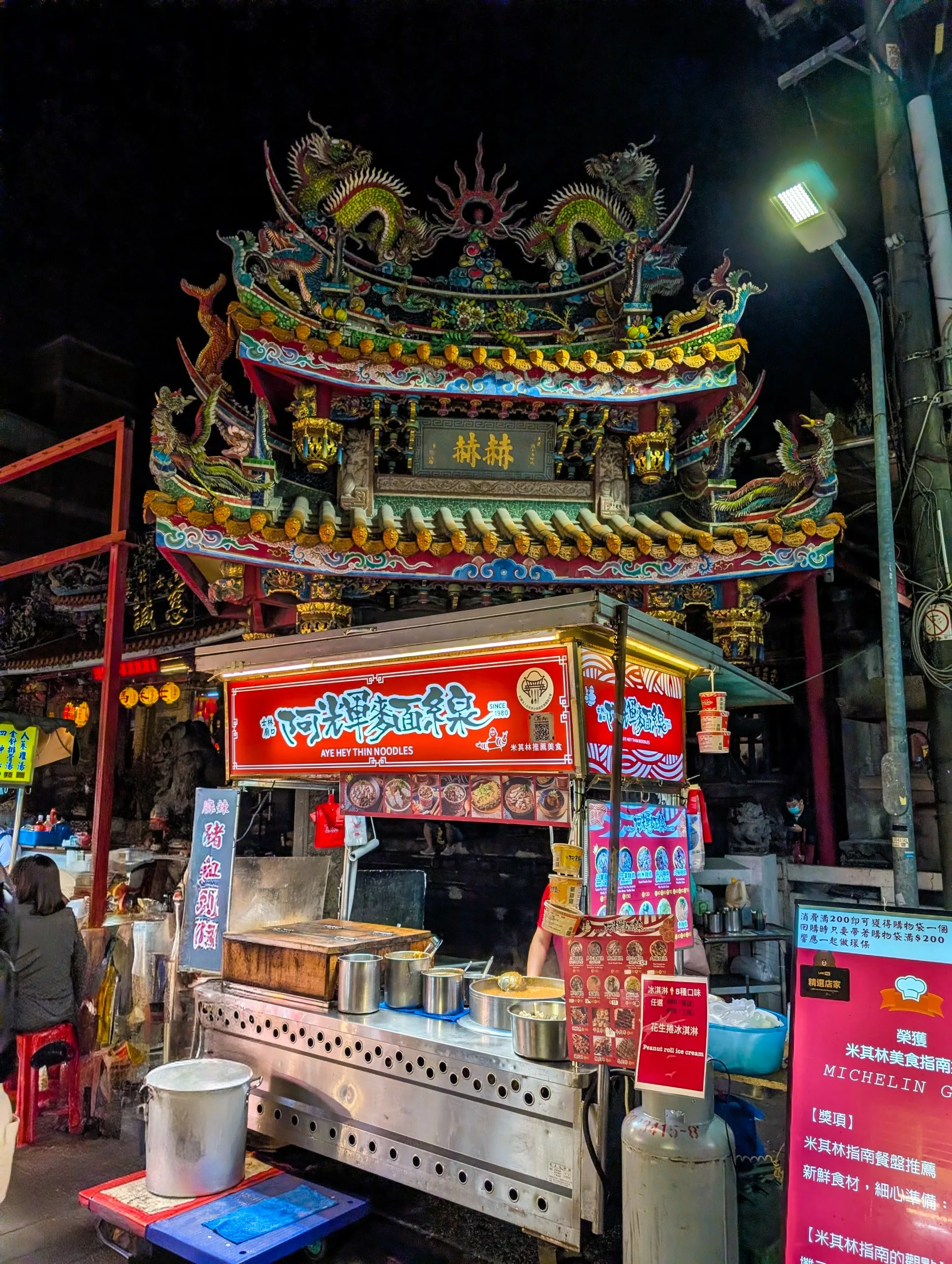 Un étal de nourriture de rue éclairé est installé devant un temple chinois orné et coloré la nuit
