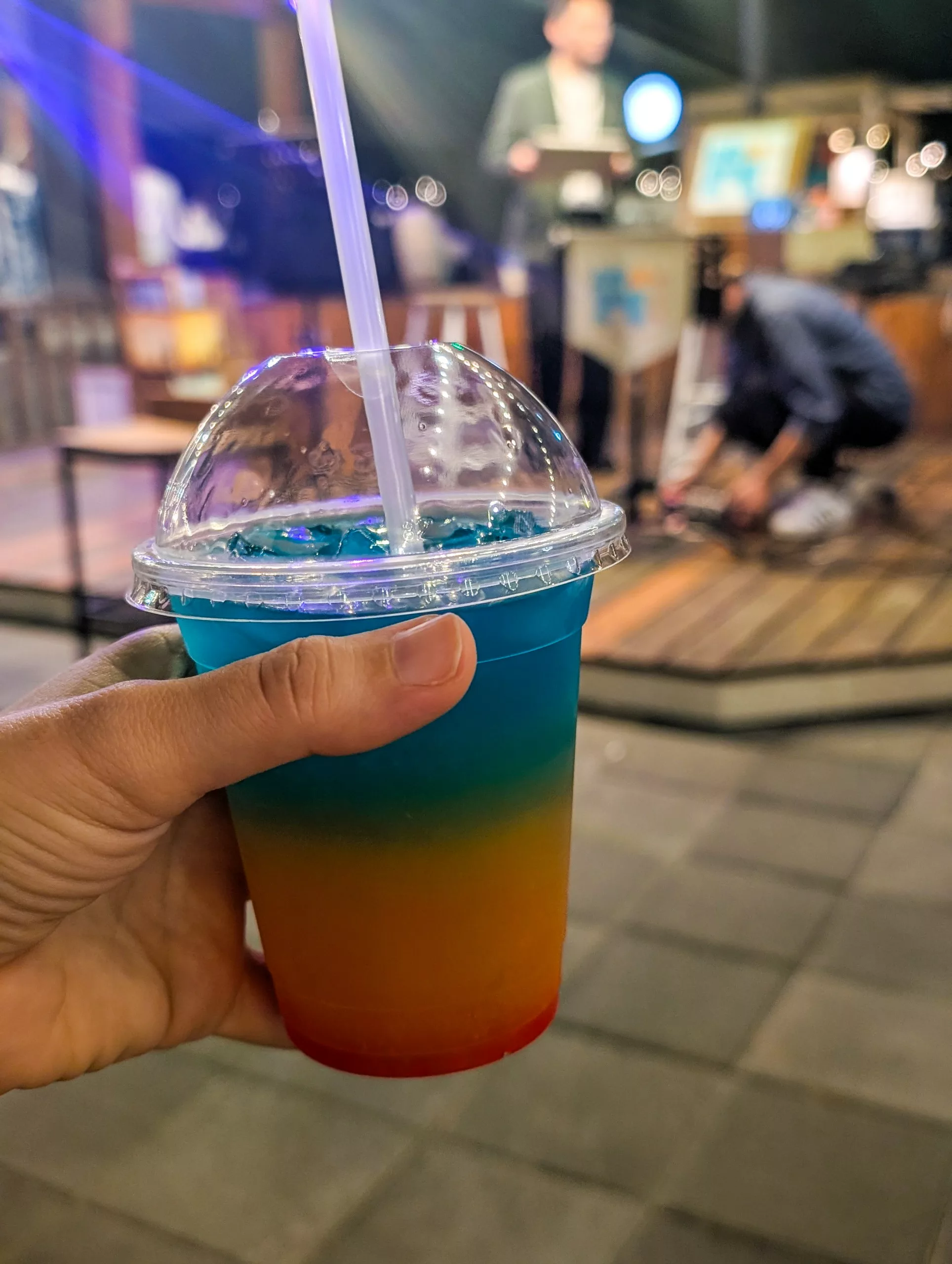 Une main tient un gobelet en plastique contenant une boisson à plusieurs couleurs, comme un arc-en-ciel.