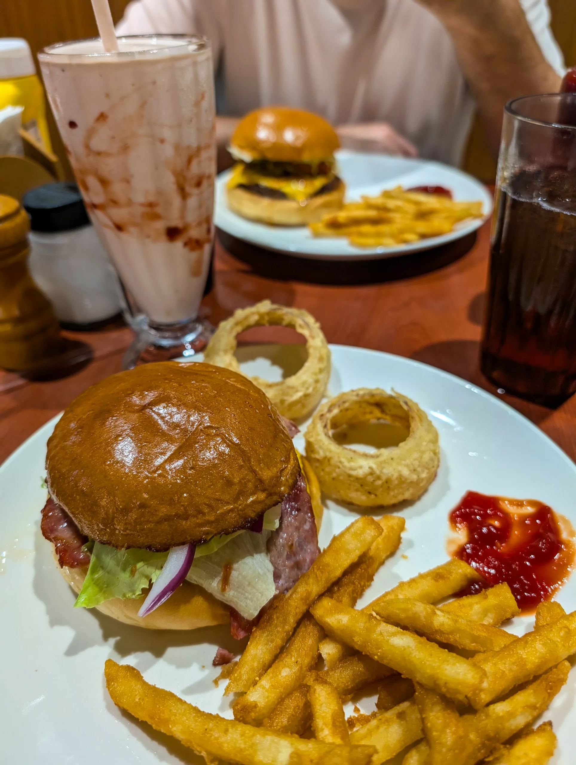 Une assiette contenant un hamburger, des rondelles d'oignon frits, des frites et du ketchup est posée à côté d'un milk-shake au chocolat. À l'arrière-plan, un autre hamburger et un soda attendent que quelqu'un déguste son repas.