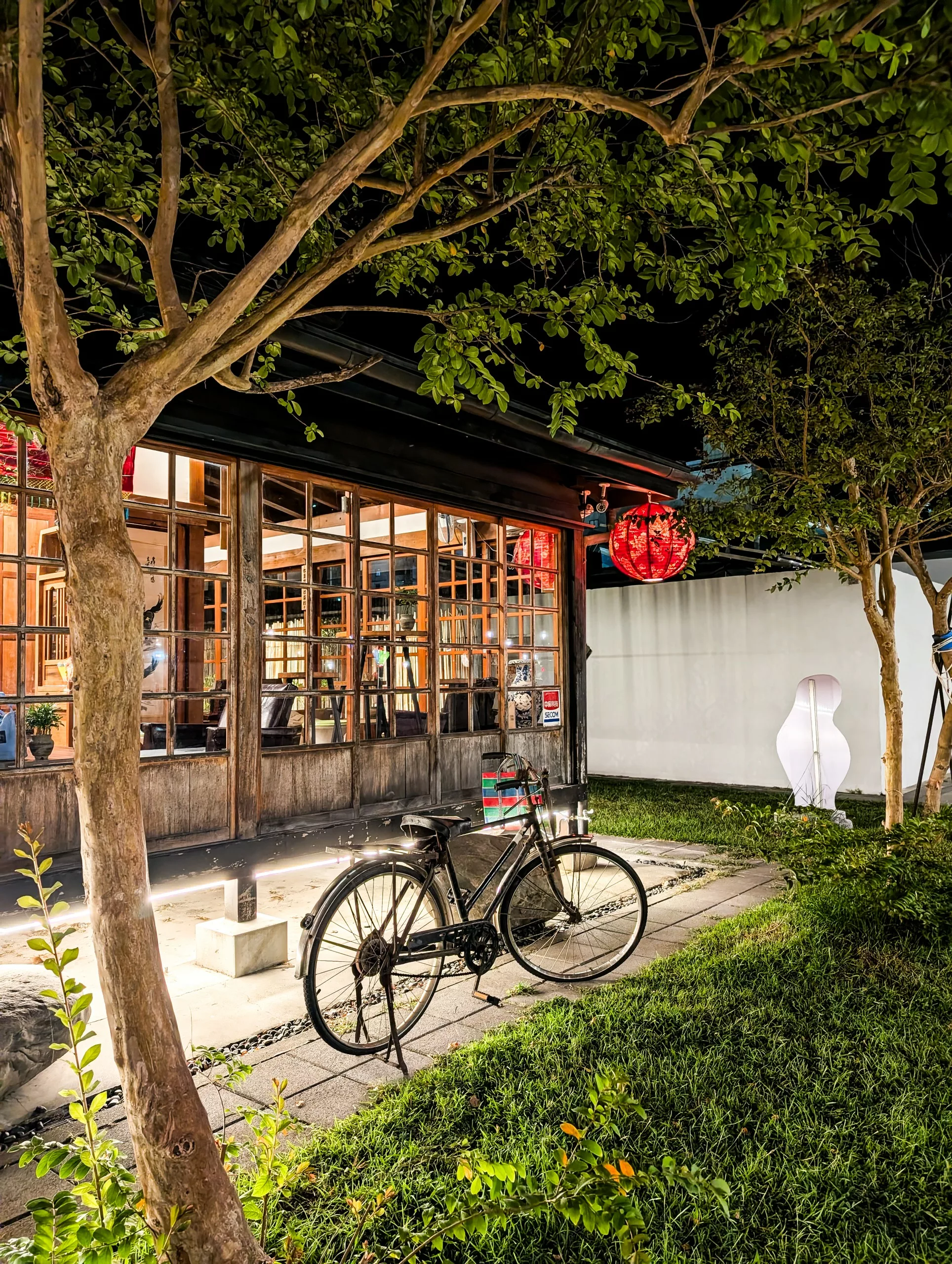 Un vélo noir est garé sur un chemin de pierre devant un bâtiment aux grandes fenêtres en bois, illuminé la nuit