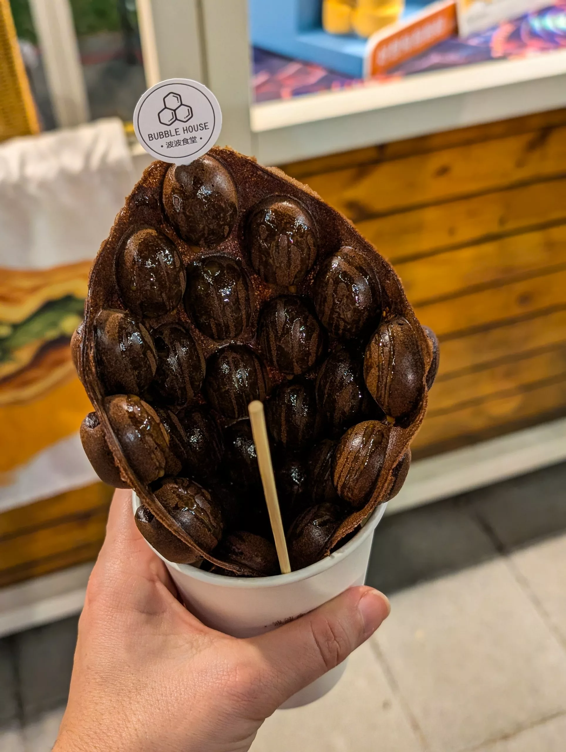 Une main tient une tasse avec une gaufre au chocolat et deux bâtonnets en bois, devant un comptoir en bois.