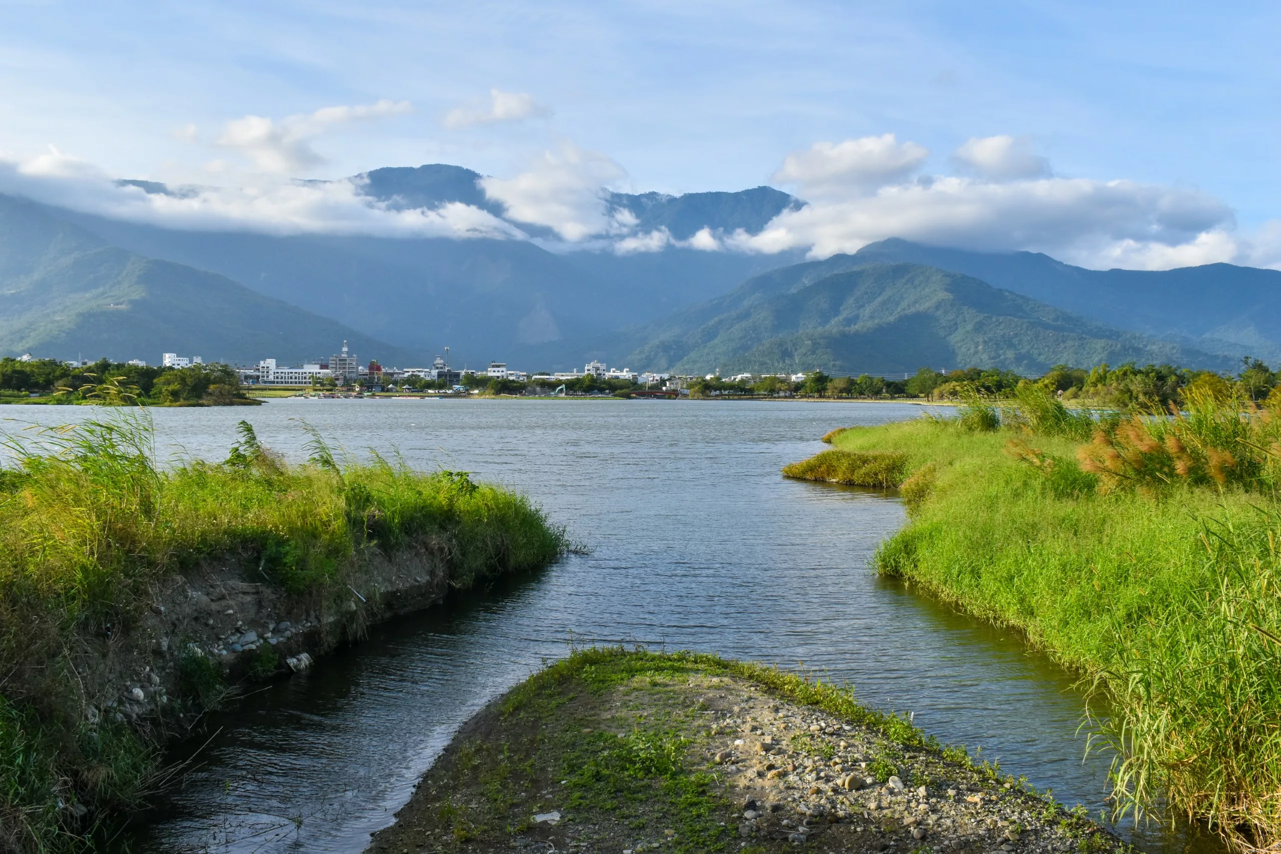 Une rivière aux rives herbeuses s'écoule vers une petite ville au loin, entourée de montagnes boisées et de nuages dans un ciel bleu