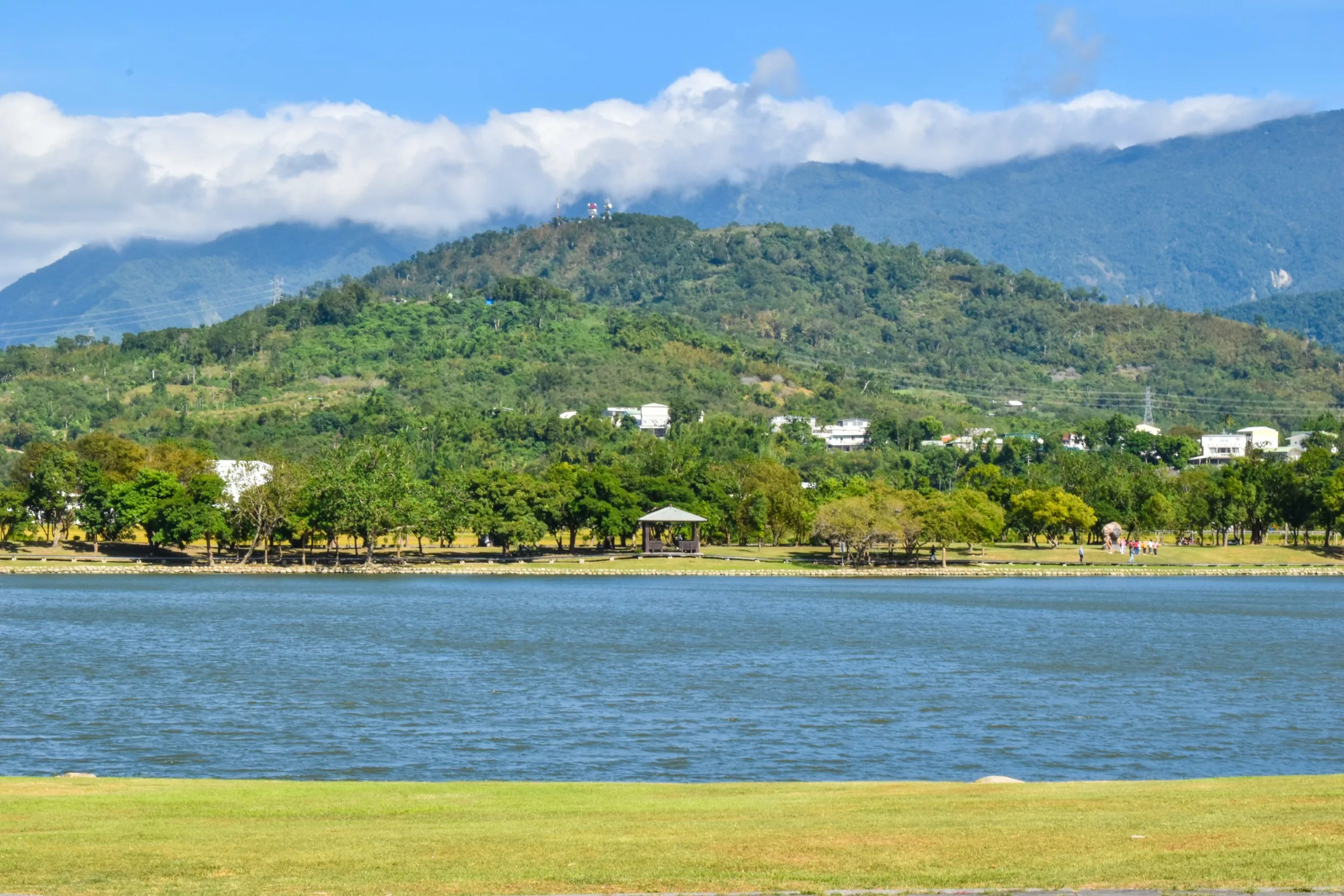 Un lac calme bordé d'herbe verte, avec un belvédère et des arbres en arrière-plan, et des collines boisées