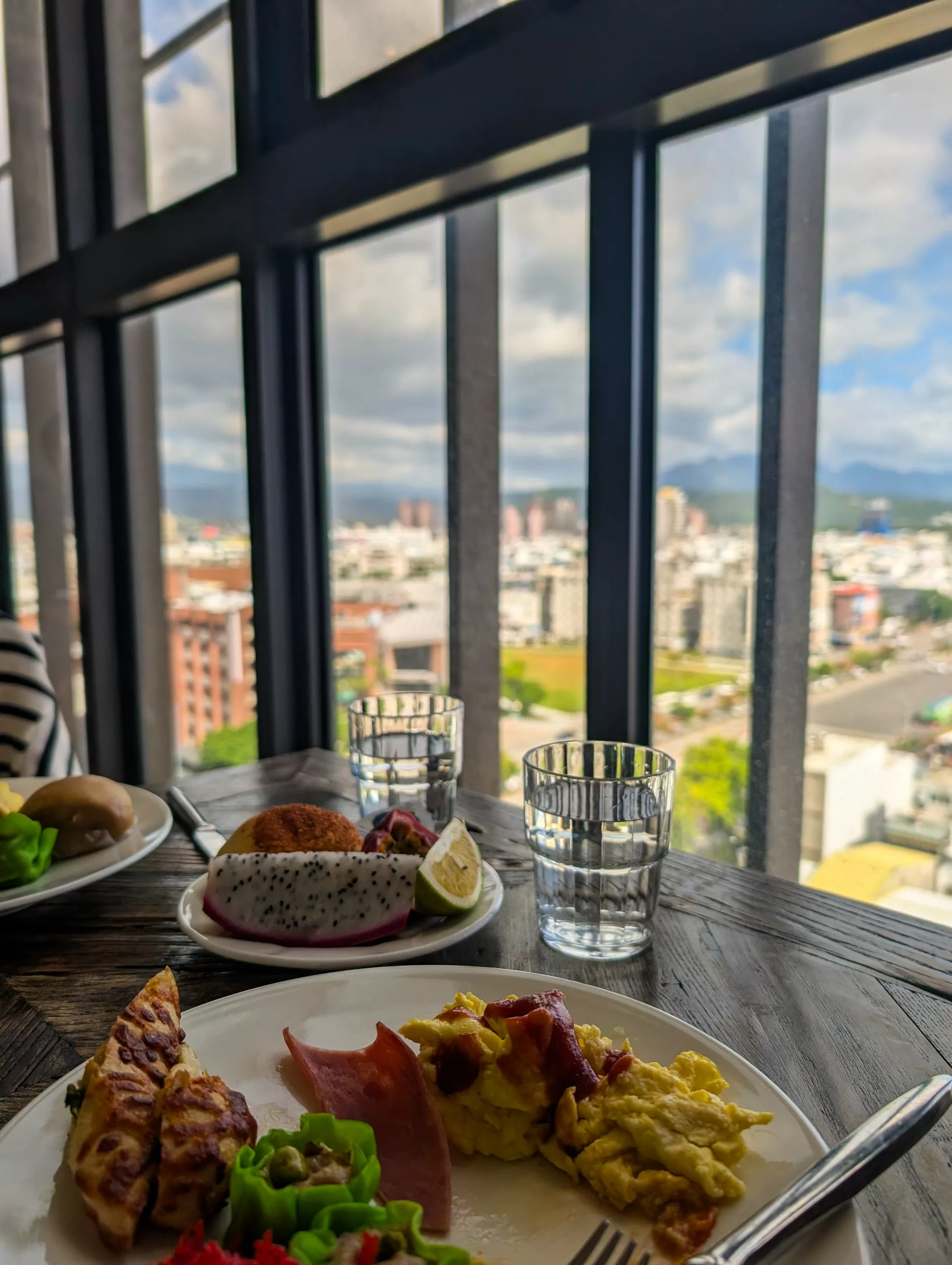 Des assiettes de petit-déjeuner contenant des œufs brouillés, du jambon, des toasts, des légumes et des fruits du dragon sont posées sur une table en bois près d'une grande fenêtre.
