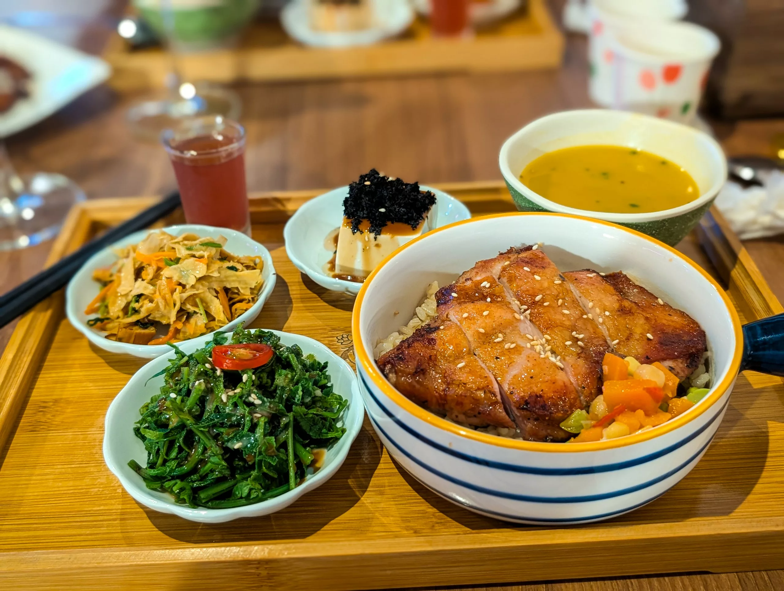 Un plateau en bois contient un bol de poulet grillé sur du riz avec des légumes, une soupe, des légumes verts marinés, des légumes verts sautés, du tofu avec une garniture noire et un petit verre de liquide rouge