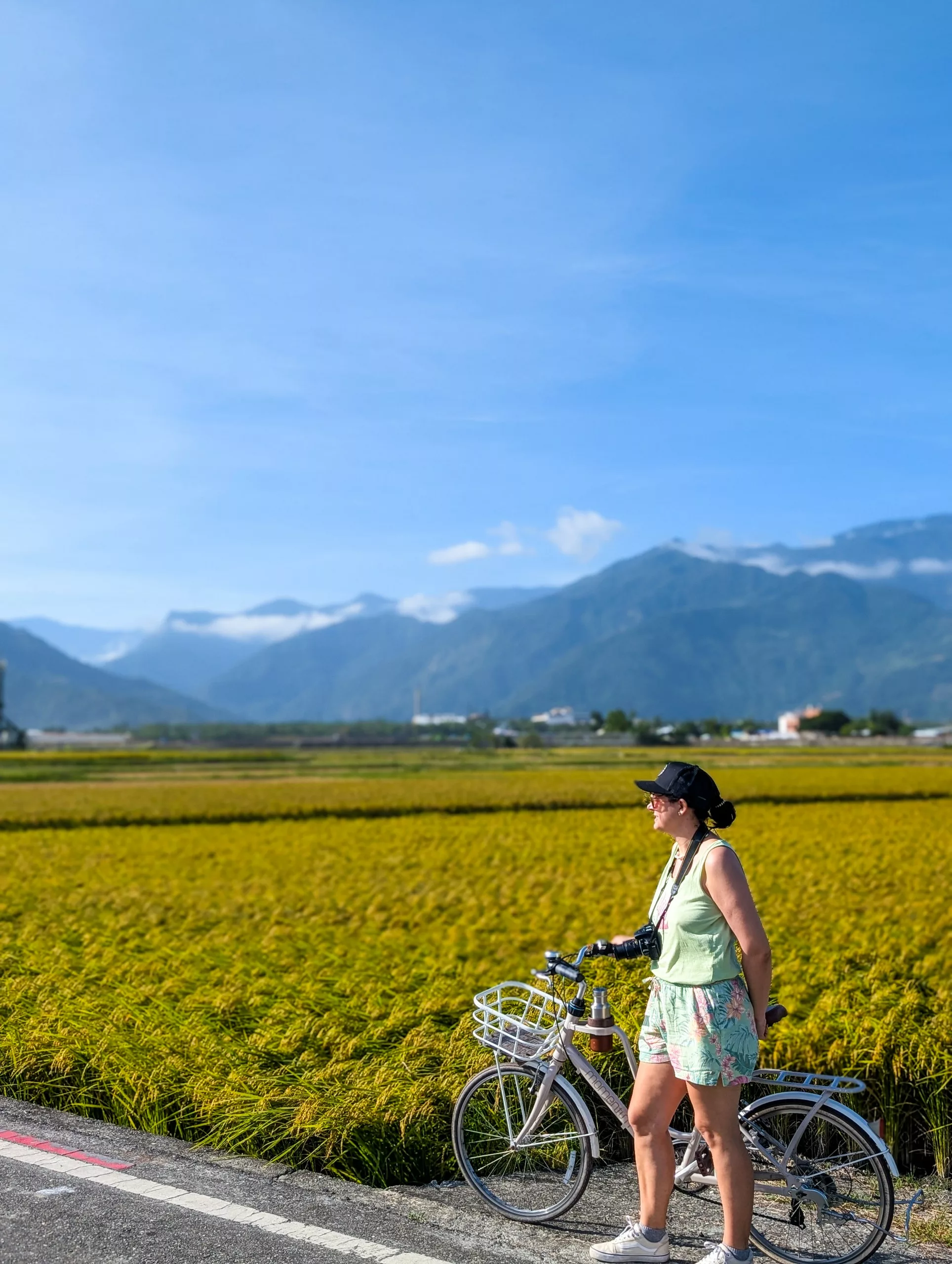Un femme portant une casquette se tient avec un vélo devant des cultures jaunes, avec des montagnes lointaines et un ciel bleu derrière.
