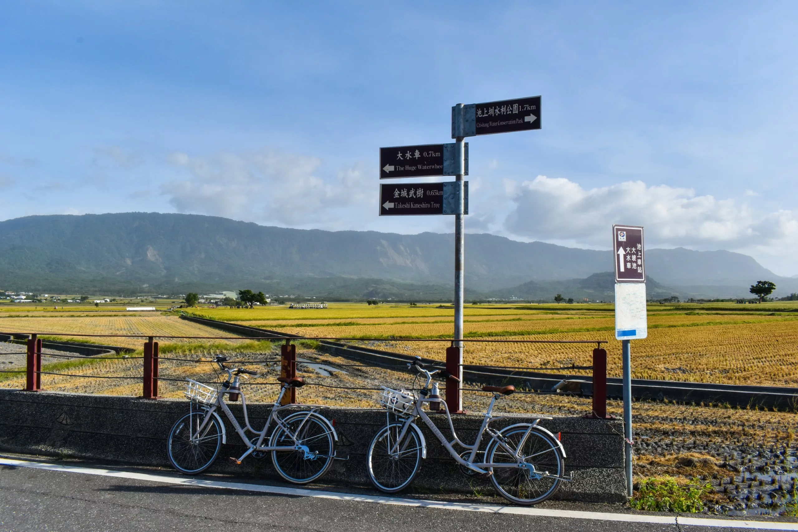 Deux bicyclettes sont garées près d'un muret de pierre, au bord d'une route de campagne.