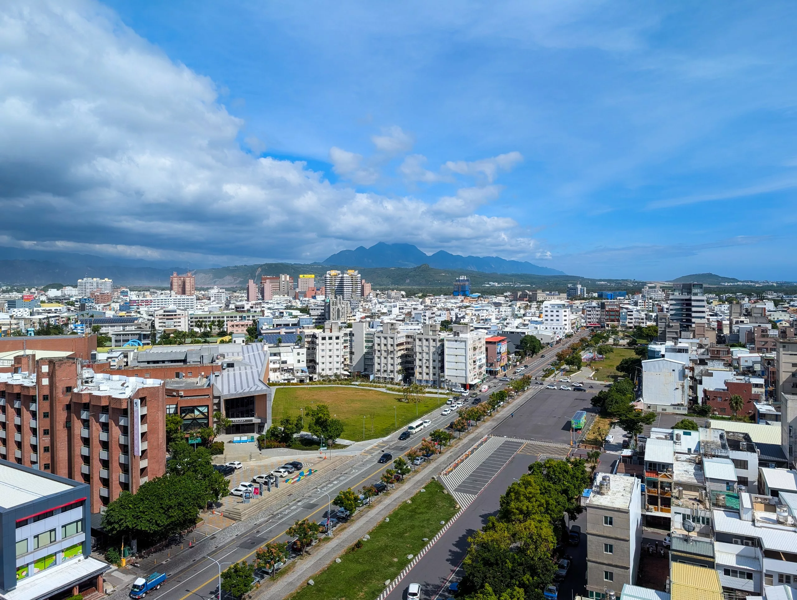 Un paysage urbain composé d'immeubles de moyenne hauteur, d'une large route et de voitures garées, avec de la verdure. Des montagnes lointaines s'élèvent sous un ciel bleu parsemé de nuages.