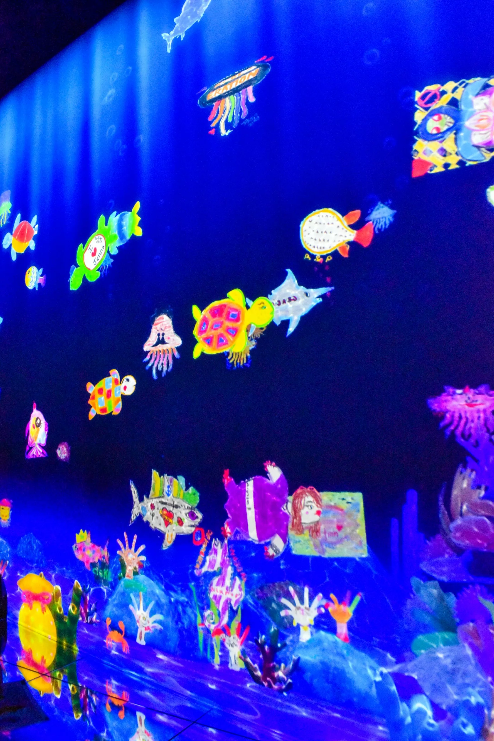 Une pièce sombre présente une scène sous-marine très éclairée avec des créatures marines colorées ressemblant à des dessins animés. Une expérience qui rappelle les expositions d'art numérique et qui est incontournable pour tous ceux qui se demandent que faire à Tokyo.