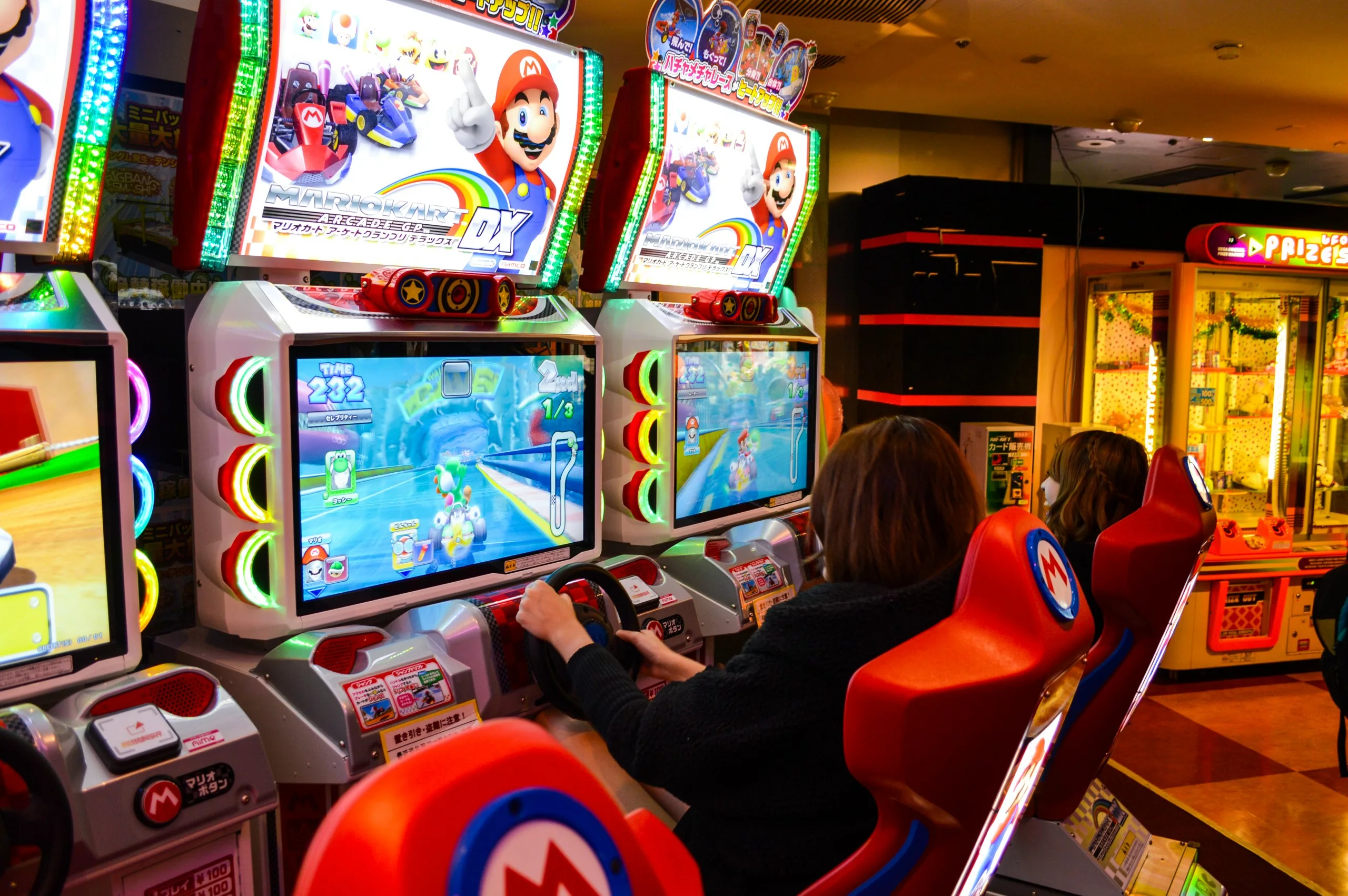 Deux personnes sont assises dans des sièges d'arcade de course rouges sur le thème de Mario, et jouent à un jeu de conduite sur de grands écrans