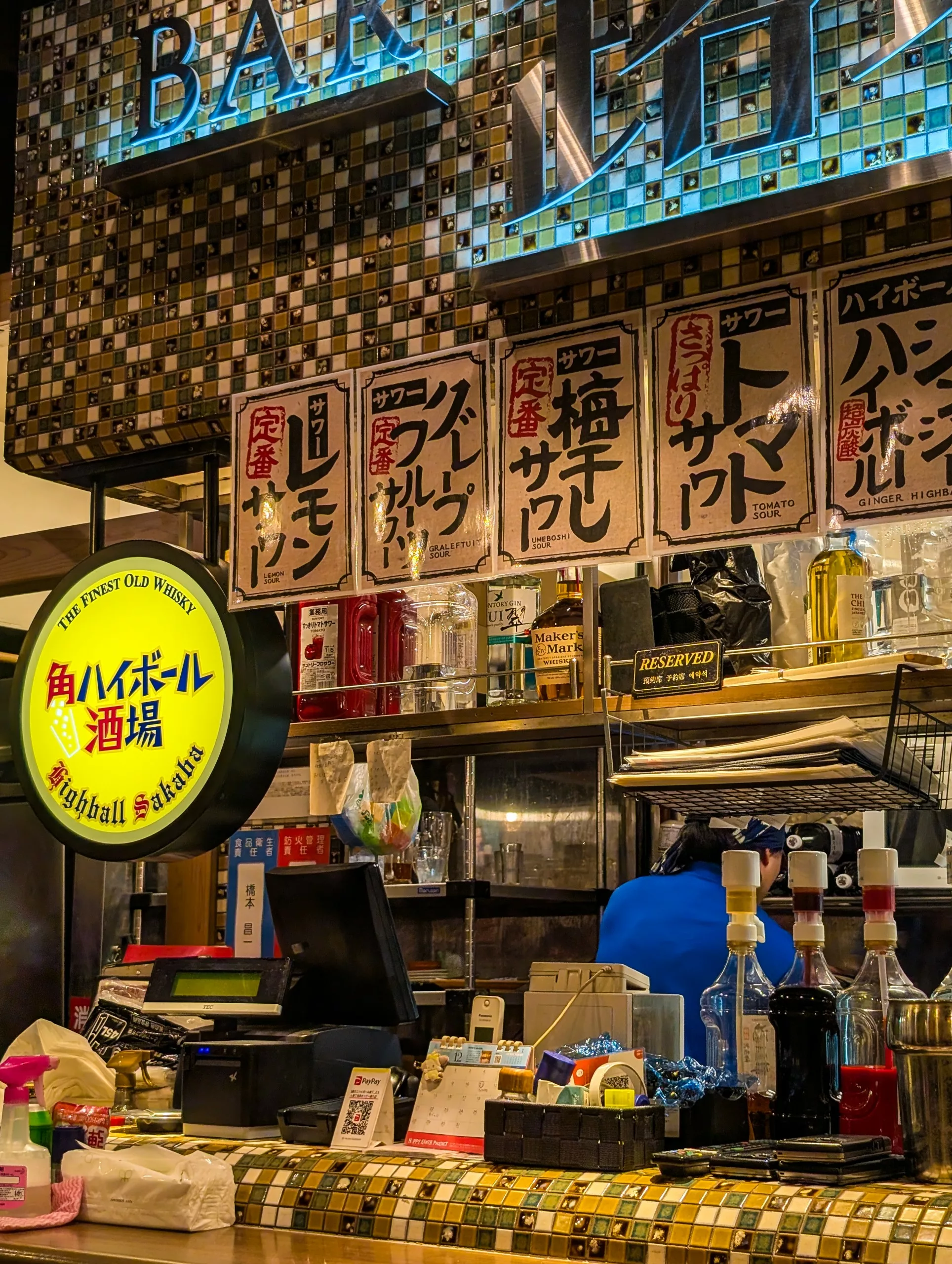 Un comptoir de bar avec diverses bouteilles, une caisse enregistreuse et des distributeurs de sirop. Des menus manuscrits en japonais sont accrochés au-dessus. Un néon rond annonce des highballs - un endroit à ne pas manquer si vous vous demandez que faire à Tokyo.