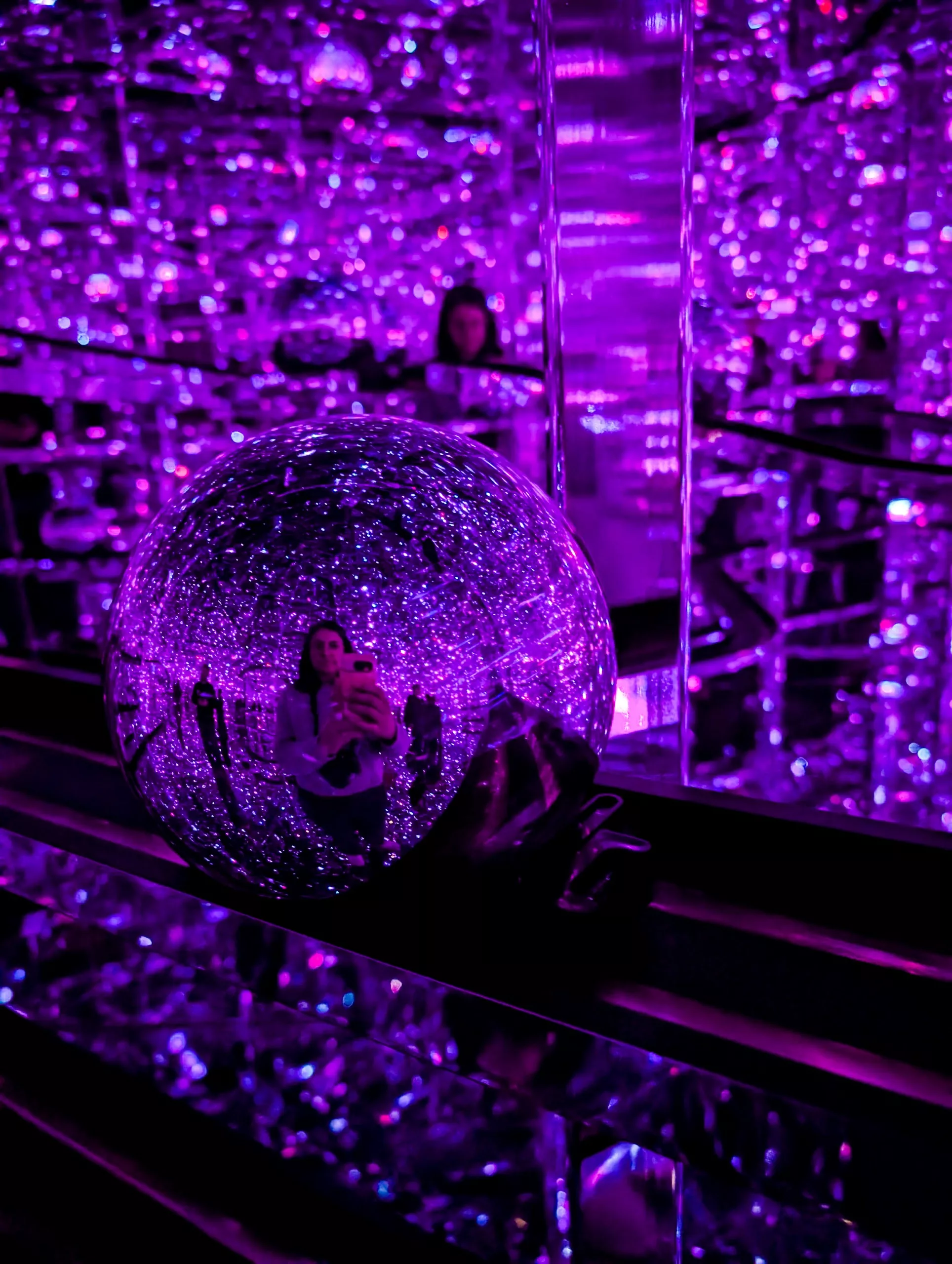 Une personne se reflète plusieurs fois dans des miroirs et dans une grande sphère réfléchissante, baignée de lumières violettes pour un effet kaléidoscopique - un endroit à ne pas manquer si vous cherchez que faire à Tokyo. La personne immortalise le moment avec son téléphone.