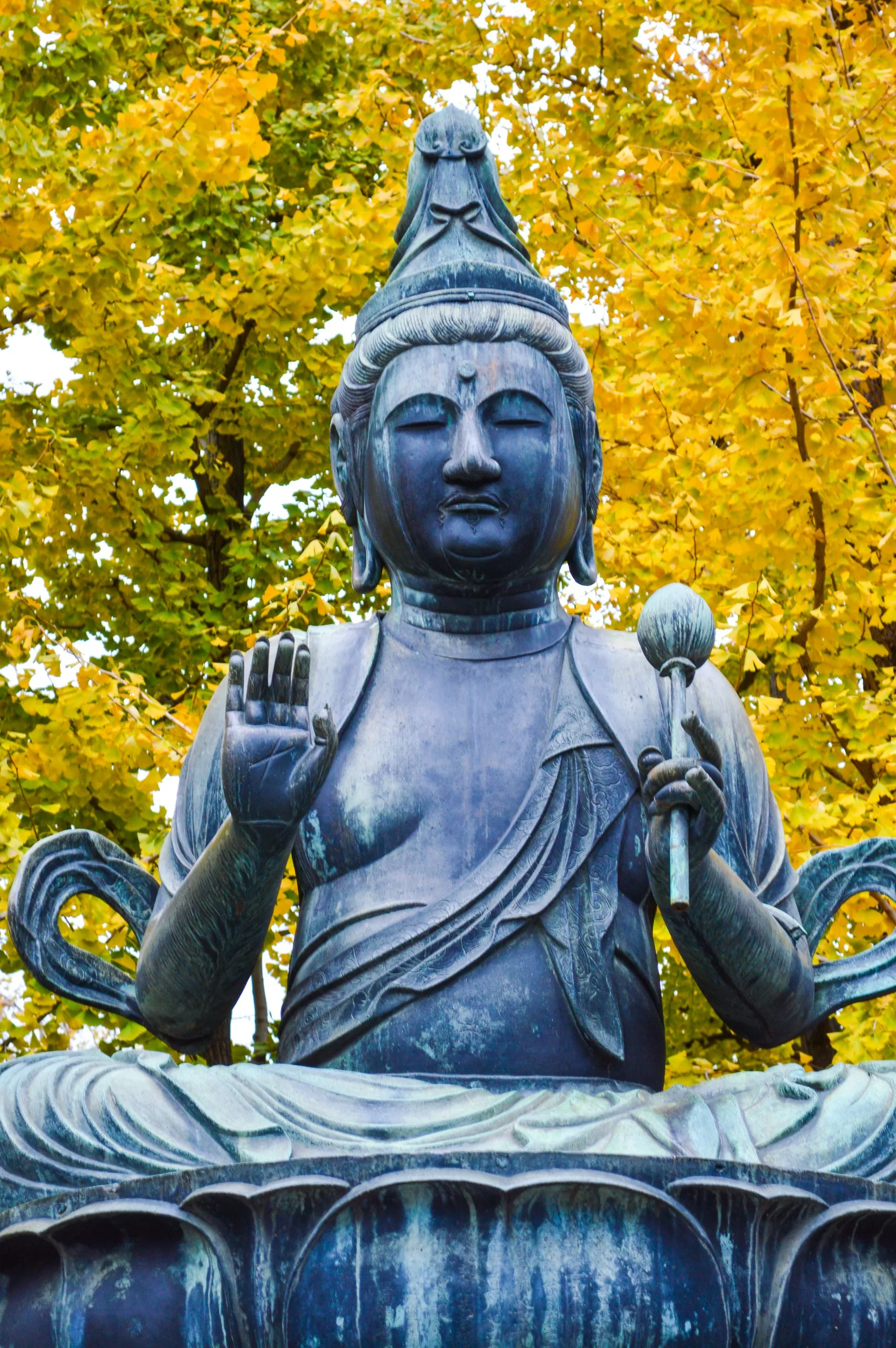 Une statue de bronze représentant un personnage assis en tenue traditionnelle, une main levée et l'autre tenant un objet - un spectacle inspirant pour tous ceux qui cherchent que faire à Tokyo au milieu des feuilles d'automne d'un jaune éclatant.