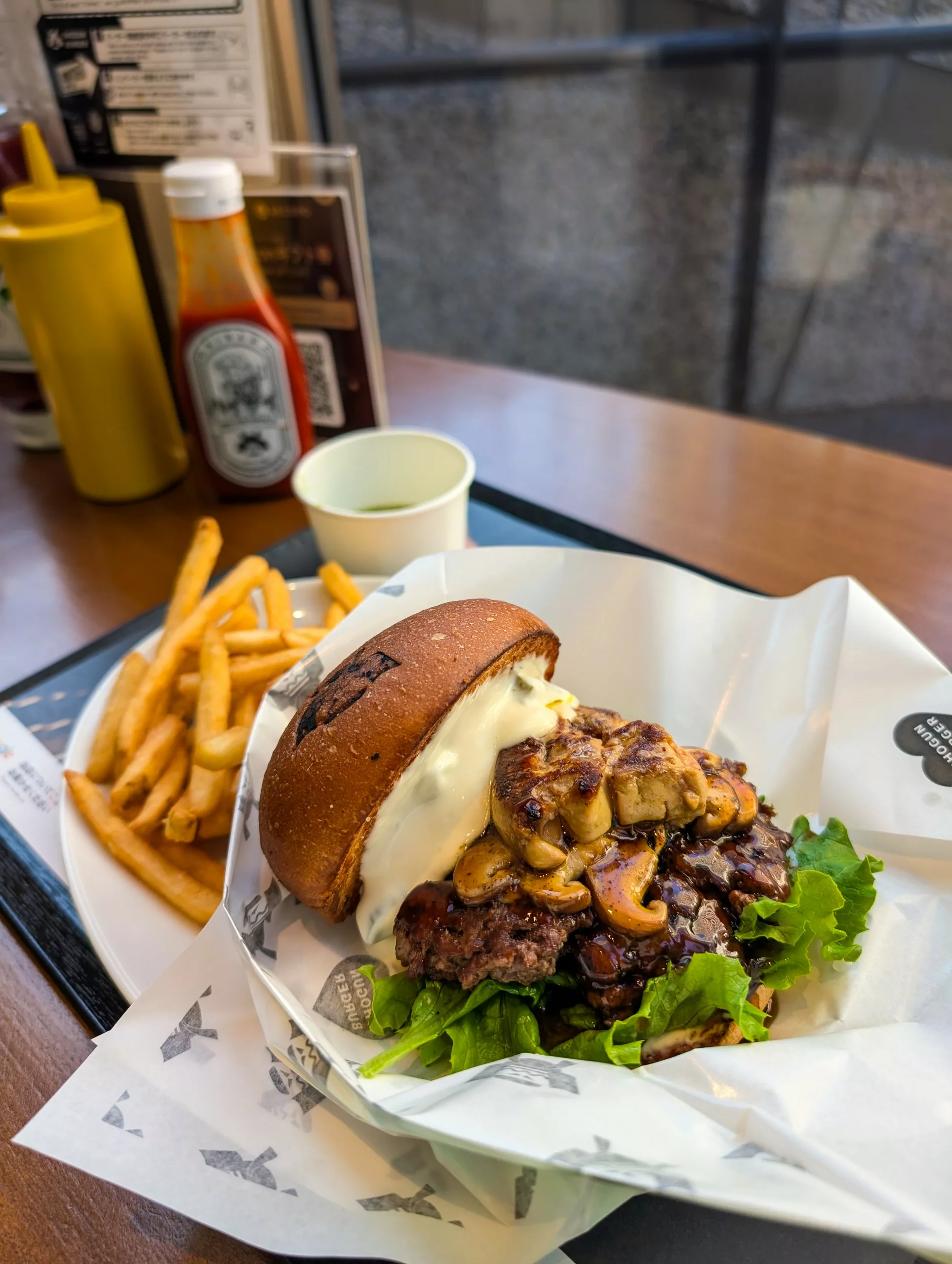 Un hamburger avec de la laitue, des champignons sautés, de la sauce et un petit pain brun est servi avec des frites - le carburant idéal pour préparer votre liste de choses à faire à Tokyo. Des bouteilles de ketchup et de moutarde ainsi qu'un gobelet de boisson sont posés sur la table en bois.