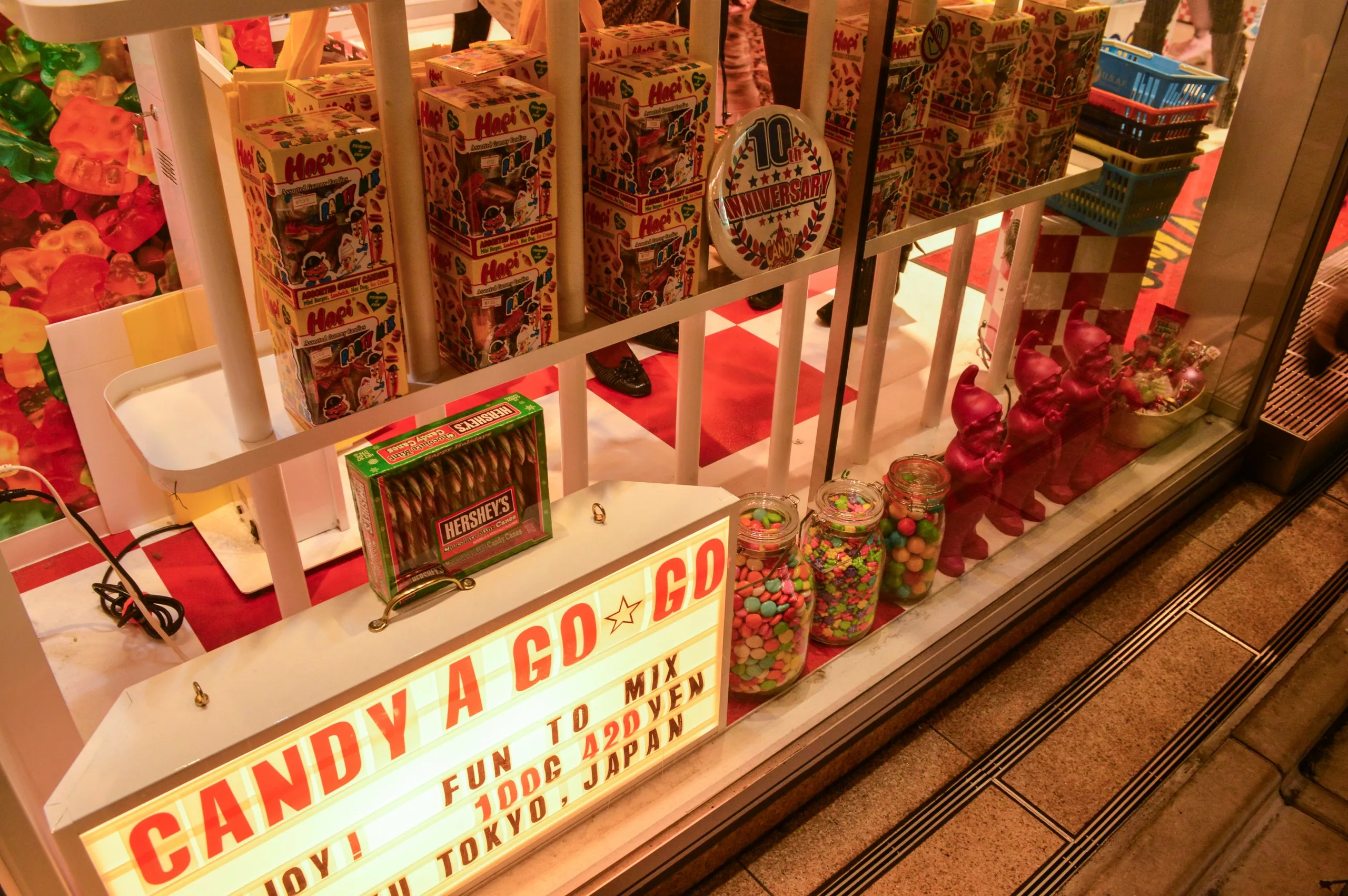 Une vitrine présente des bonbonnières colorées, des bocaux remplis de boules de gomme et du chocolat Hershey's. Le nom "CANDY A GO GO" brille au-dessus, célébrant le 10e anniversaire de la boutique. La mention "CANDY A GO GO" brille au-dessus de la vitrine, célébrant le 10e anniversaire de la boutique. Une halte sucrée pour tous ceux qui cherchent à savoir ce qu'il faut faire à Tokyo.