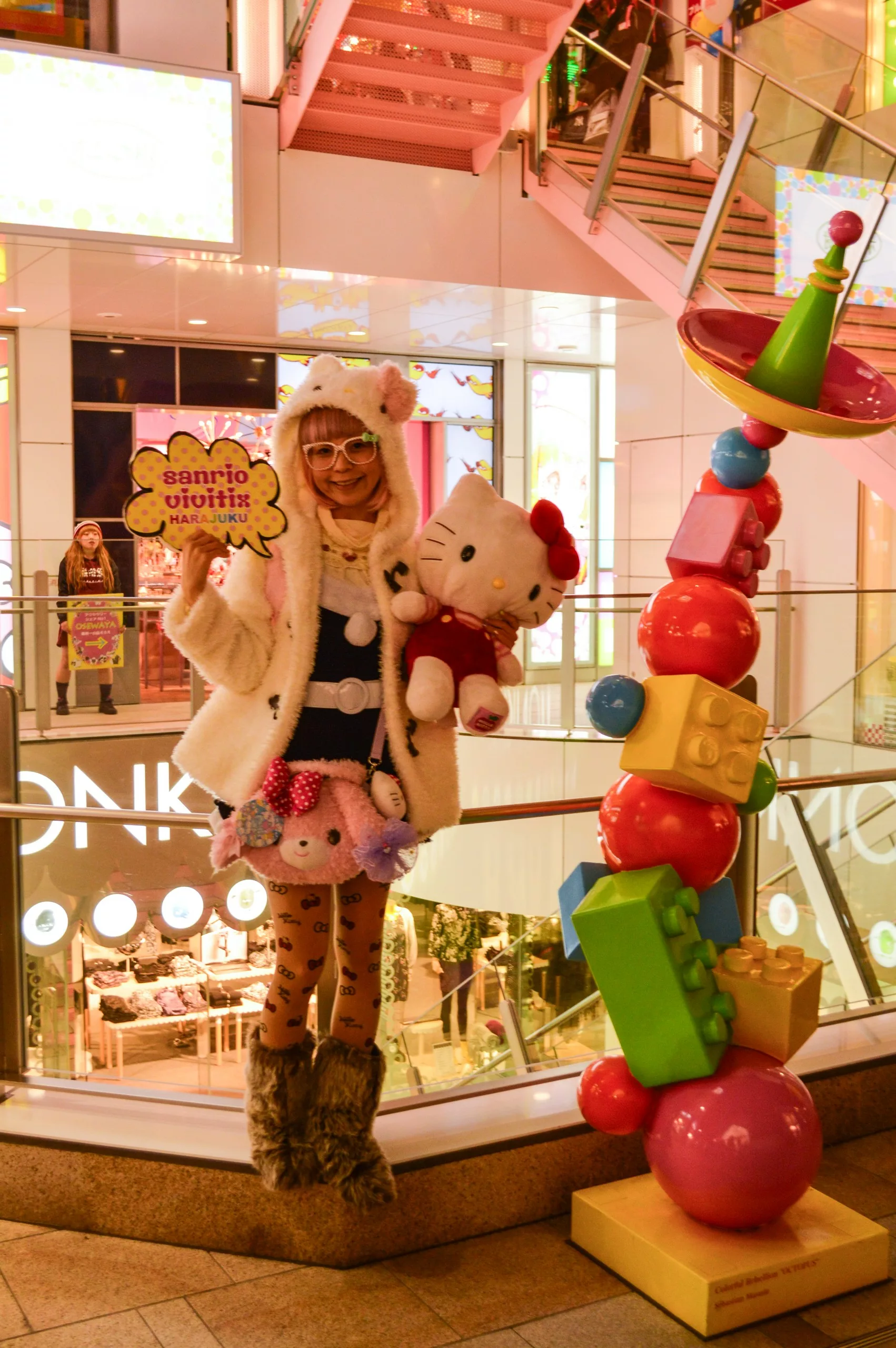 Une personne vêtue d'un manteau à capuche en fourrure blanche, d'une robe à pois et de collants sur le thème des animaux tient une peluche Hello Kitty et un panneau Sanrio - présentant les principales choses à faire pour ceux qui se demandent que faire à Tokyo- à l'intérieur d'un centre commercial animé.