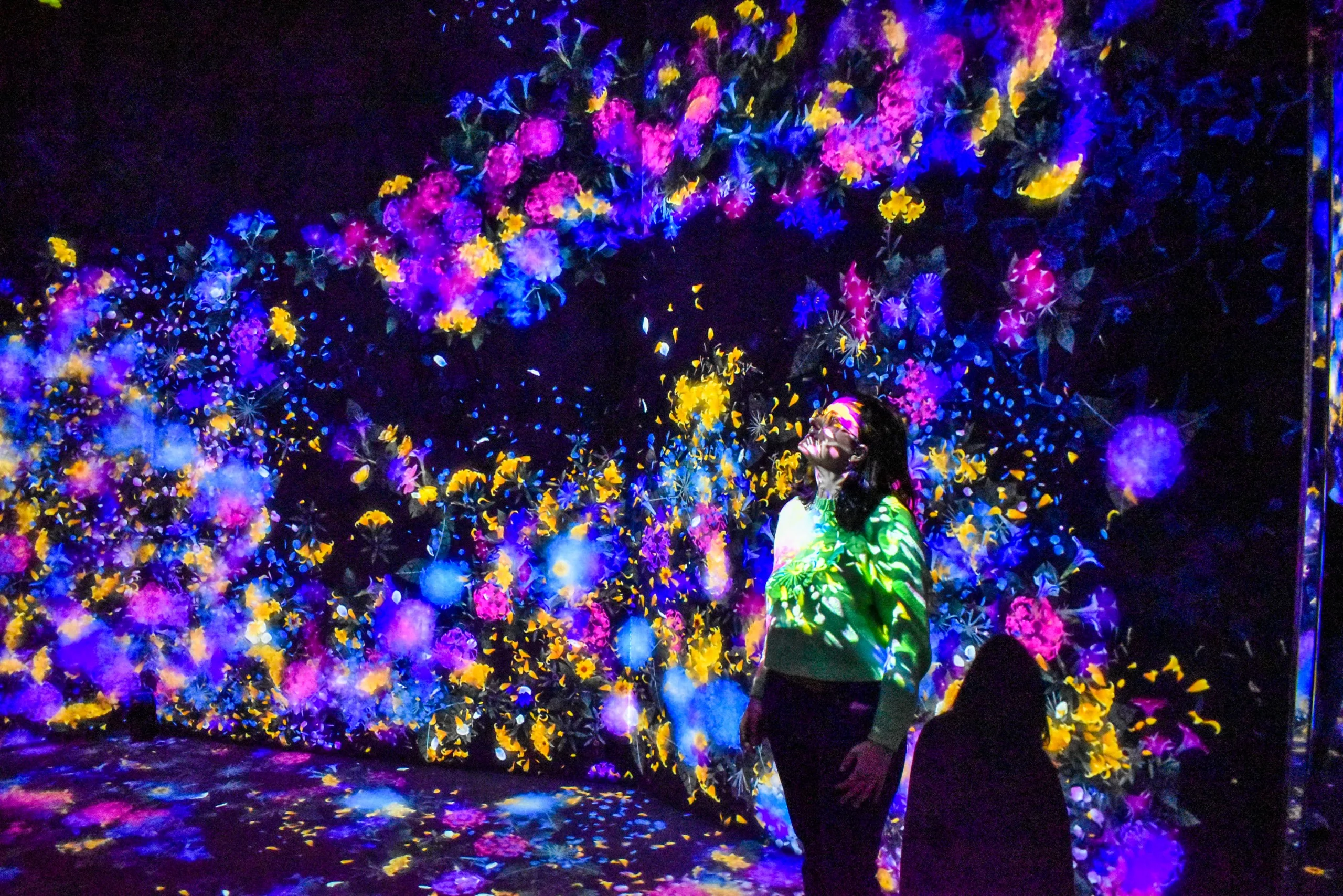 Une personne se tient devant une installation d'art numérique - l'une des expériences à ne pas manquer pour qui veut découvrir que faire à Tokyo- avec des fleurs aux couleurs vives et des motifs abstraits projetés sur un mur sombre.