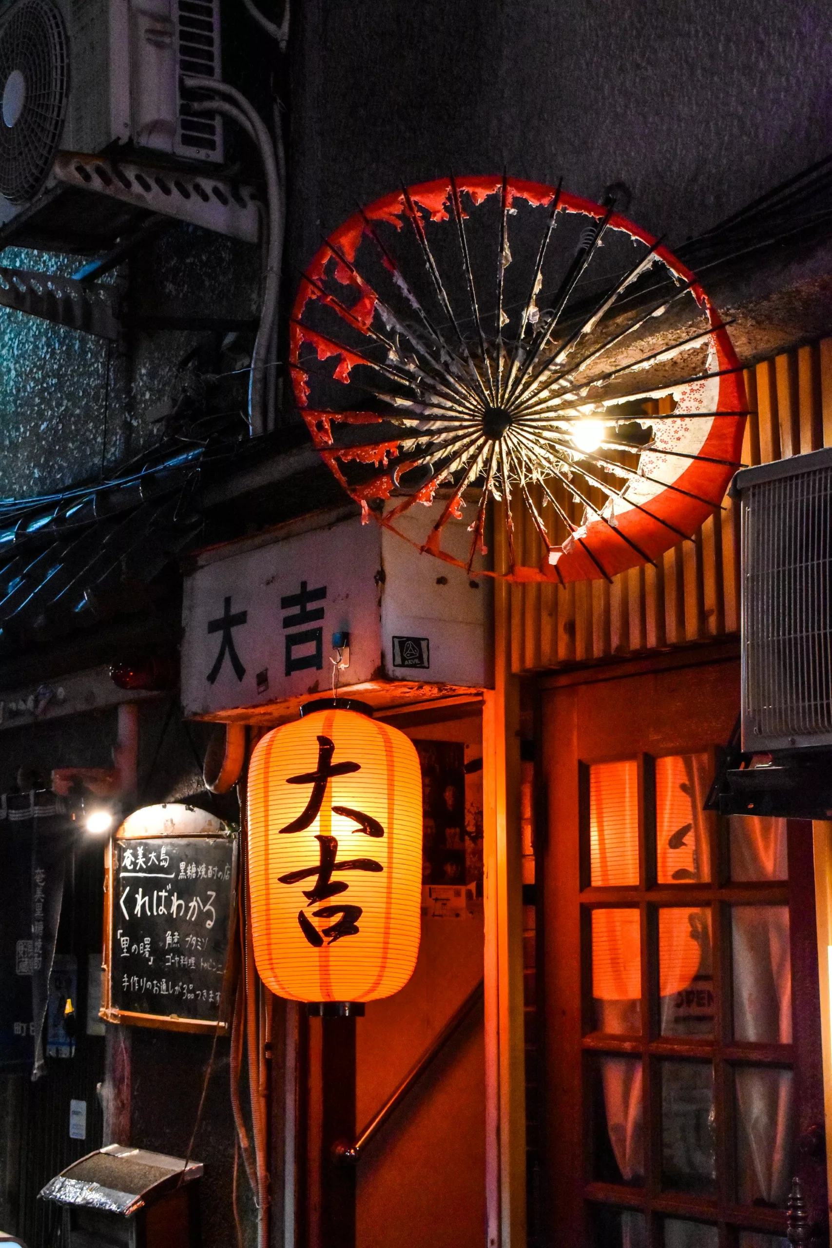 Une scène de rue japonaise chaleureusement éclairée la nuit, avec une lanterne en papier rouge, un grand parapluie décoratif accroché au mur et un panneau à la craie