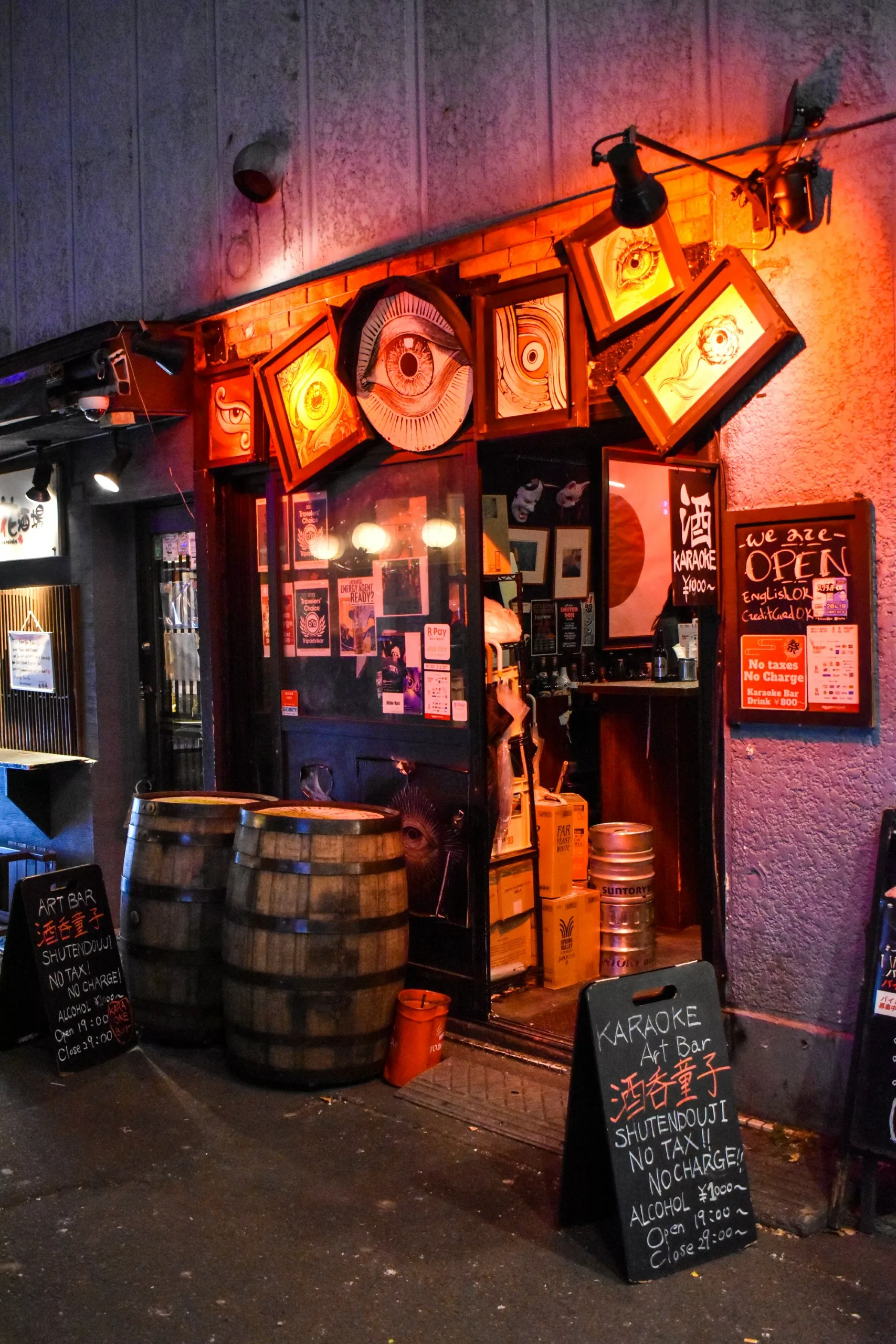 Une entrée de bar avec des tonneaux en bois à l'extérieur, un éclairage orange, des œuvres d'art encadrées en angle, des panneaux en anglais et en japonais